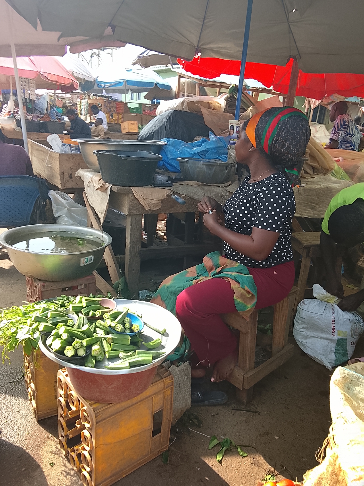 This is a medium shot of an outdoor market scene in Akwanga, Nigeria. A woman wearing a black and white polka dot shirt, red pants, and a colorful headscarf sits on a wooden stool. She is facing right, with her hands clasped in her lap. In front of her, on a table, are several bowls and crates. A large metal basin filled with water is to the left, and a pile of okra is in a large circular platter to the front. To the right of the woman, another person in a green shirt is visible from the waist down, appearing to be sorting through a white sack. Several other market stalls and shoppers are visible in the background, mostly out of focus, under various tarps and umbrellas. The ground is dirt, and the scene is illuminated by bright sunlight.