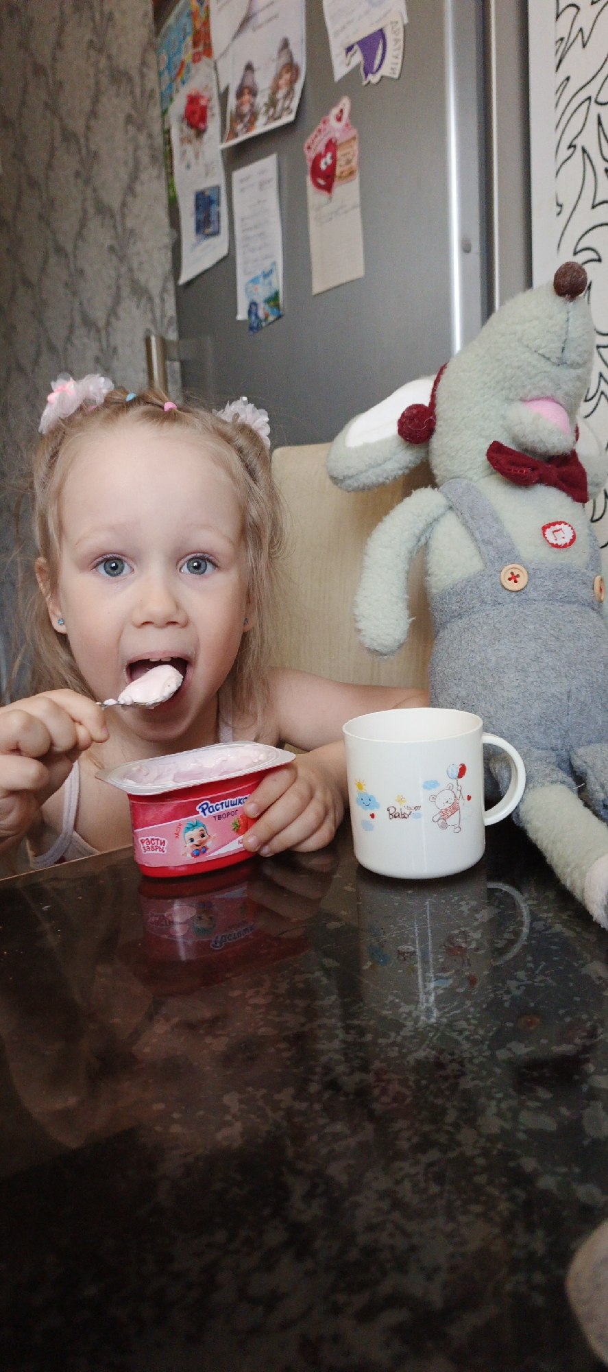 In a close-up, eye-level shot set in Chaikovsky, Russia, a young blonde child with blue eyes and hair styled in two small buns with pink hair ties is captured mid-meal. The child's mouth is open, receiving a spoonful of light pink yogurt, with an engaged and focused expression.
To the child's left, on a dark, reflective table, sits an opened container of pink yogurt. Visible Cyrillic text on the container reads "РАСТИШКА ТВОРОГ" (Rastishka Tvorog), alongside an illustration of a baby next to a strawberry. To the child's right, a white plastic mug with a handle displays the text "Happy Baby" and features cheerful illustrations of a teddy bear holding balloons and smiling clouds.
Beside the child, to their right, a large grey stuffed animal, resembling a mouse or rabbit, sits upright. It wears grey overalls with wooden buttons and a red bow tie. The background reveals a kitchen or dining area during the daytime. A large, stainless steel refrigerator is adorned with various papers, drawings, and notes, including one heart-shaped cutout and some illegible Cyrillic text; faint letters "ВКИТИ" are visible on one paper. To the left of the fridge, a light-colored wallpaper with a subtle pattern covers the wall.