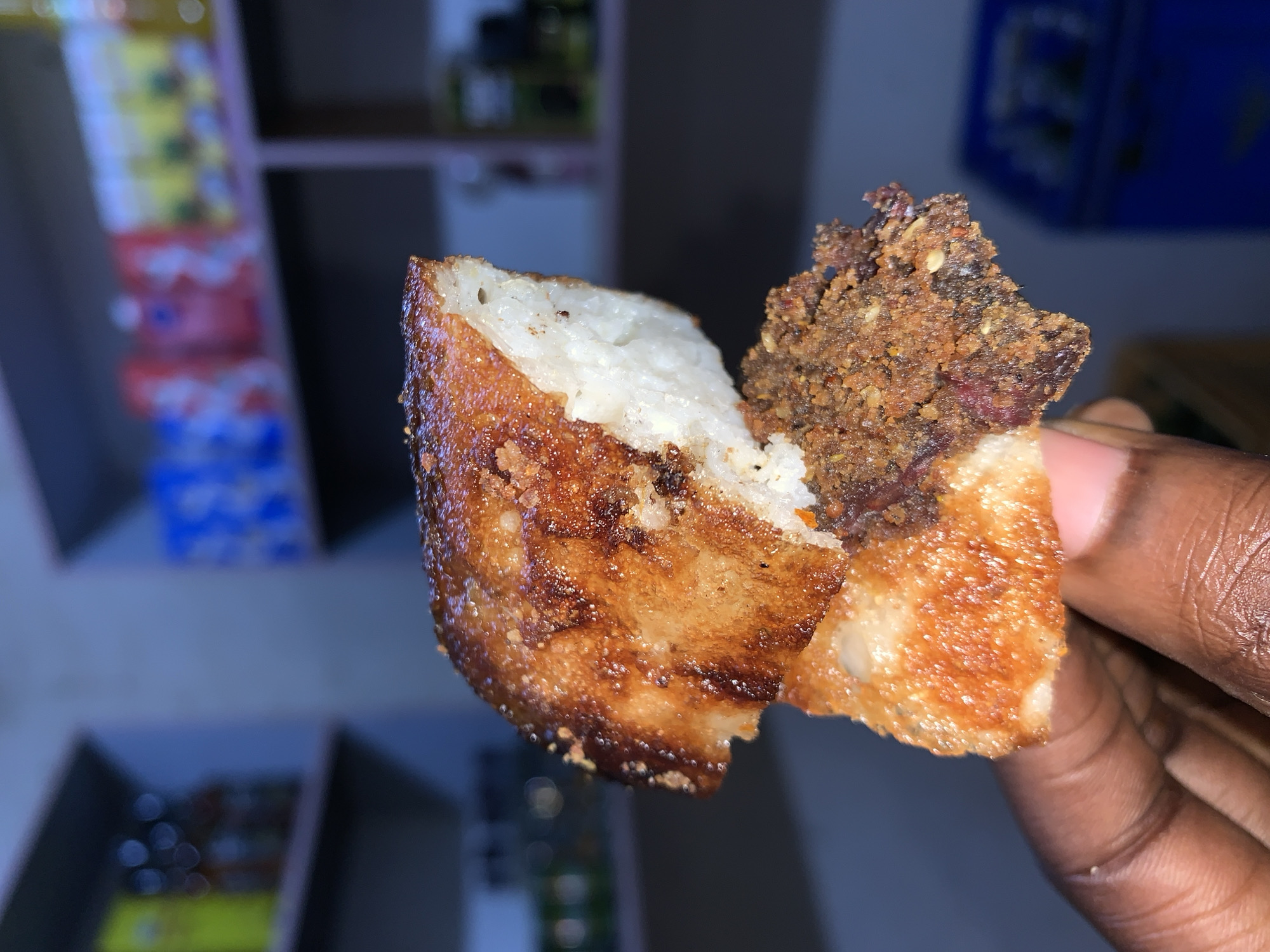 A person is holding a broken piece of what appears to be fried yam cake. The yam cake has a fried, golden-brown exterior with a white, porous interior. It is filled with a dark brown, crumbly filling. The person holding the cake has dark skin. The background is blurred, but shows a shelf with various items. The location is suggested to be near the New Stadium in Nigeria.