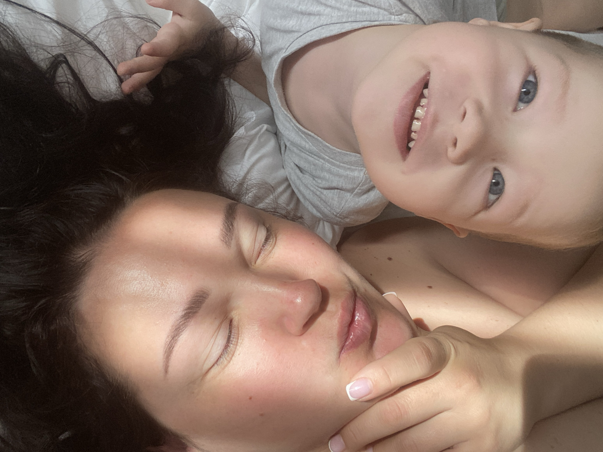 A woman and child are lying together in what appears to be a bed. The woman has dark hair and closed eyes, and she is touching her face with her hand. The child, who is wearing a gray shirt, is smiling and looking at the camera. The scene is brightly lit, suggesting daylight. The location is not discernible from the image.