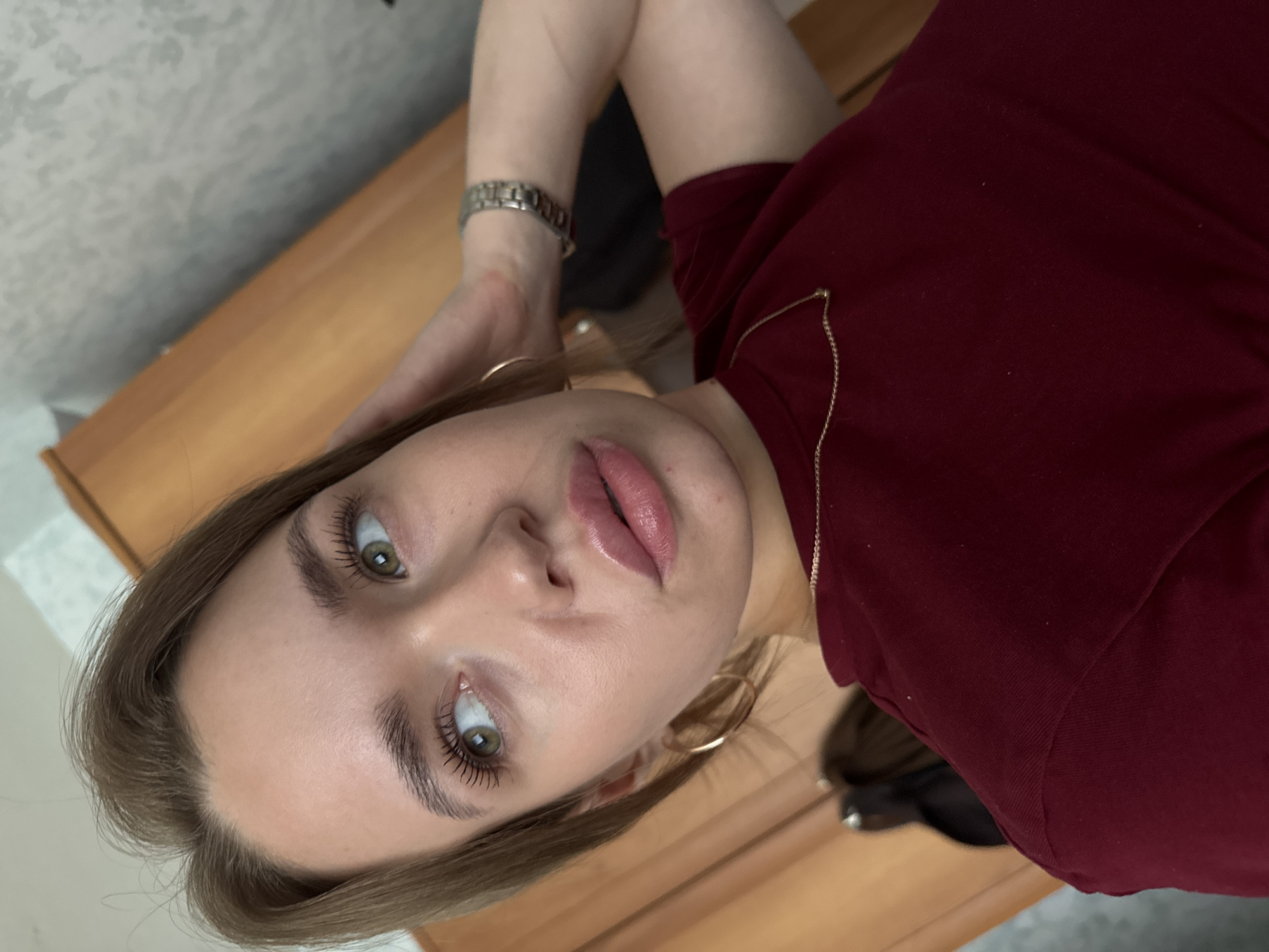 The media file contains a close-up, overhead shot of a young woman lying down. She is wearing a dark red top and a silver watch on her left wrist. She has long, light brown hair and is wearing hoop earrings. Her makeup includes mascara and lip gloss. The background appears to be a wooden surface, possibly a desk or table, with a textured, light-colored wall behind it. The lighting suggests an indoor setting, likely during the daytime. The woman's eyes are open and she is looking towards the camera. There is no visible text in the image. The woman appears relaxed, with one arm resting behind her head. The overall impression is a casual, personal photograph.