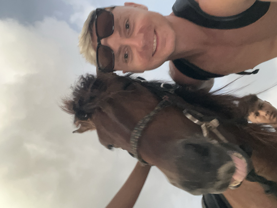 The image displays a low-angle selfie taken in Jimbaran, Indonesia, featuring a man, a horse, and a second individual against a cloudy sky. In the foreground, on the right side, a fair-skinned male with short, blonde hair and dark sunglasses pushed up on his head smiles towards the camera. He is shirtless, with black backpack straps visible on his shoulders. Adjacent to him on the left, the head of a brown horse occupies a significant portion of the frame. The horse is equipped with a bridle featuring a decorative, possibly braided, browband and noseband. Its mouth is slightly open, revealing a pink tongue and gums. Below the horse's head, a second male with dark hair is partially visible, looking towards the camera. The upper background consists entirely of a light gray, overcast sky with indistinct cloud formations. The primary man and the horse are in close proximity, suggesting an interactive activity.