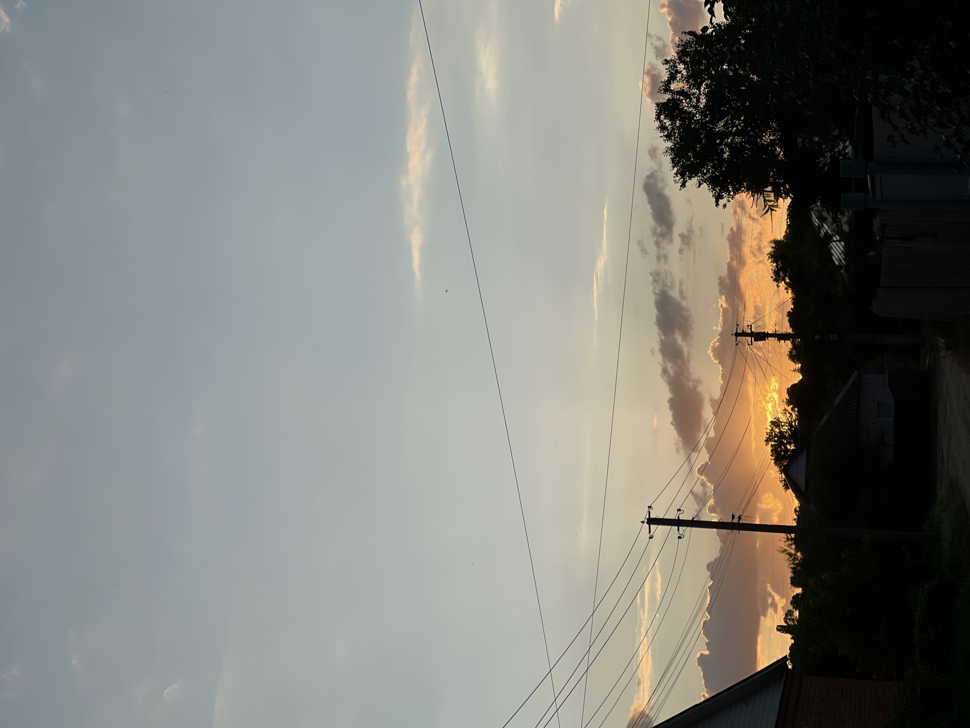 This media file captures a serene sunset scene in Krasnokutsk, Ukraine. The image is oriented vertically, with the sky occupying the left two-thirds of the frame and the lower portion revealing a rural or semi-rural setting.
The sky is a canvas of soft pastels, transitioning from a pale blue at the top to warmer hues of orange and yellow near the horizon. Wispy clouds, tinged with pink and gold by the setting sun, are scattered across the sky. A few power lines crisscross the scene, supported by utility poles, indicating the presence of infrastructure in the area. Silhouetted against the vibrant sky are trees and the dark outlines of houses or other buildings, suggesting a residential or village environment. The bright glow of the sun peeking through the clouds implies it is either just before or just after the sun has dipped below the horizon, typical of a late evening or early morning.
The overall mood is peaceful and tranquil, evoking the quiet beauty of a rural sunset. There are no people visible in the frame. The setting appears to be a common, everyday landscape, rather than a specific event. No text is visible in the image. The weather appears clear with scattered clouds, and the time of day is definitively sunset.