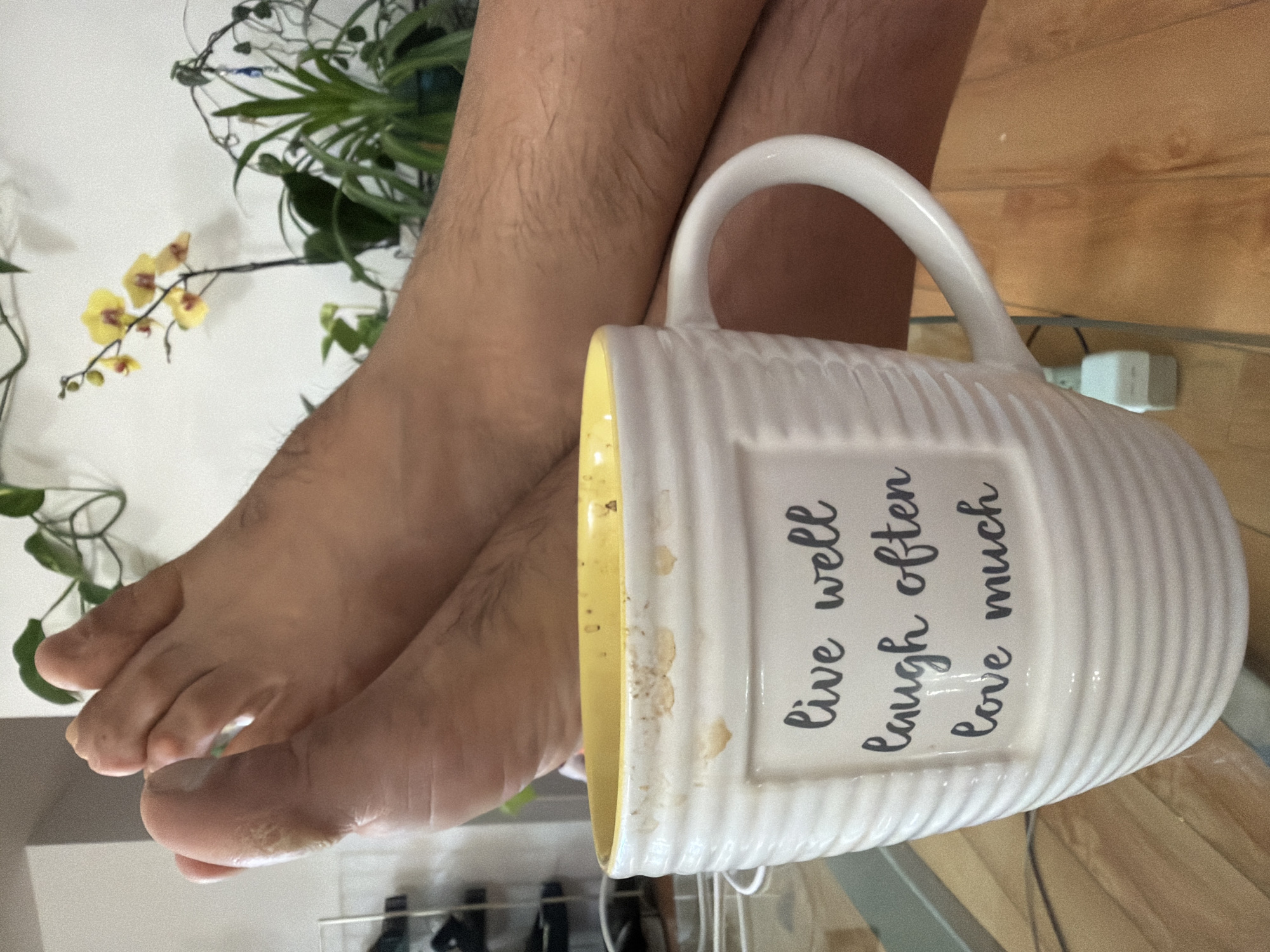 The image shows a close-up of a person's legs and feet, positioned next to a white mug with a yellow interior. The mug has a ribbed texture and is decorated with the phrase "live well laugh often love much" in black cursive lettering. The mug appears to have some stains on its rim. The person's legs are covered in hair, and their feet are visible, with toes splayed. Behind the legs and mug, a blurred background reveals houseplants, including a flowering orchid with yellow blossoms, and a wooden surface that may be a table or floor. The lighting suggests an indoor setting, possibly during the daytime. There is no indication of the weather or the specific location. The overall impression is one of a casual, relaxed moment.