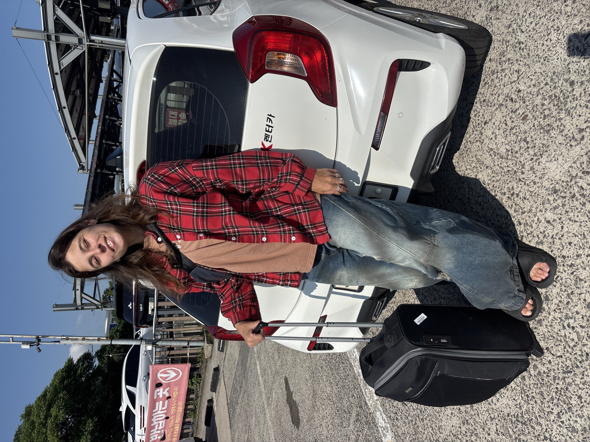 The image shows a young woman leaning against the rear of a white car in an outdoor parking lot. She is wearing a red and black plaid shirt, a brown t-shirt underneath, and distressed denim jeans. Her hair is long and brown, and she is smiling at the camera. She has rings on her fingers and is wearing black slides. Beside her is a black rolling suitcase with its handle extended.
The scene appears to be a car rental lot, indicated by the presence of multiple cars, a large banner with Korean text, and overhead structures that might be part of a parking garage or service area. The banner displays Korean characters which translate to "Return Location" and "Please organize and leave the key in the vehicle. We are not responsible for lost items." Another sign on the car's rear window also has Korean text, possibly a rental car company name or branding, and a license plate is partially visible. The sky is clear and blue, suggesting a sunny day, and shadows on the ground indicate it is likely daytime. The overall impression is of someone preparing for or concluding a car rental.
