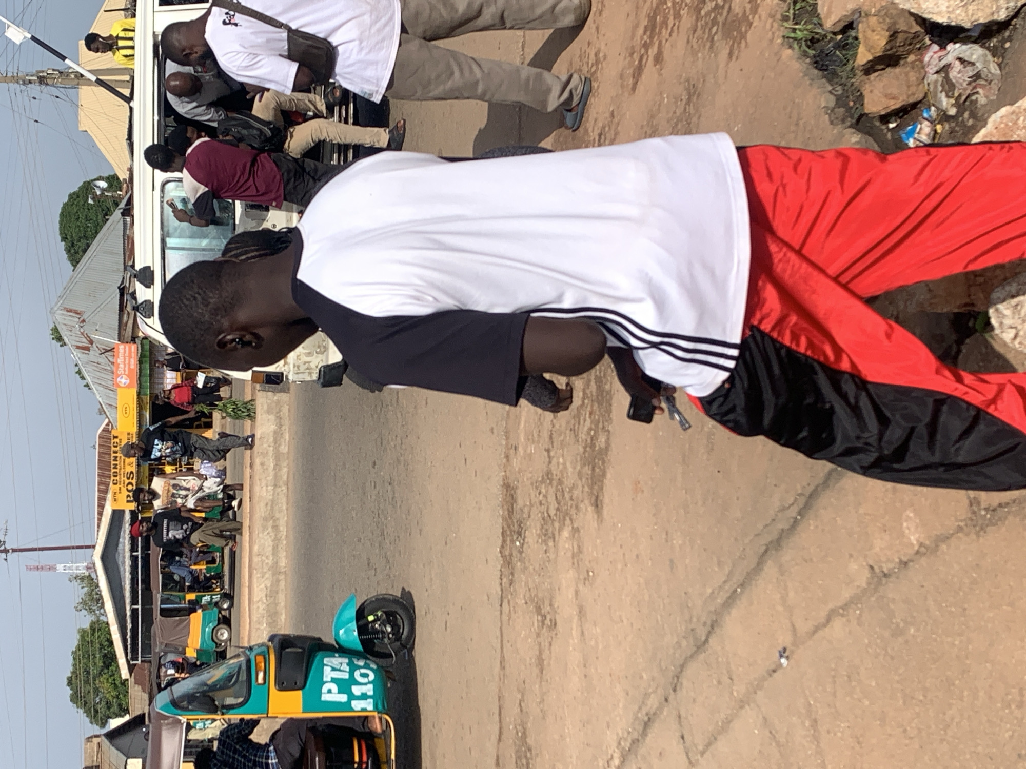 In Gura Top, Nigeria, a man in a black and white Adidas shirt and red and black athletic pants is captured from behind, walking away from the camera on a dirt road. The man holds a mobile phone in his right hand, near his waist. To his left, a turquoise and yellow tuk-tuk is parked. In the background, other vehicles, including a white van, and several people are visible near a street-side business with a red and yellow awning. The sky above is clear and blue.