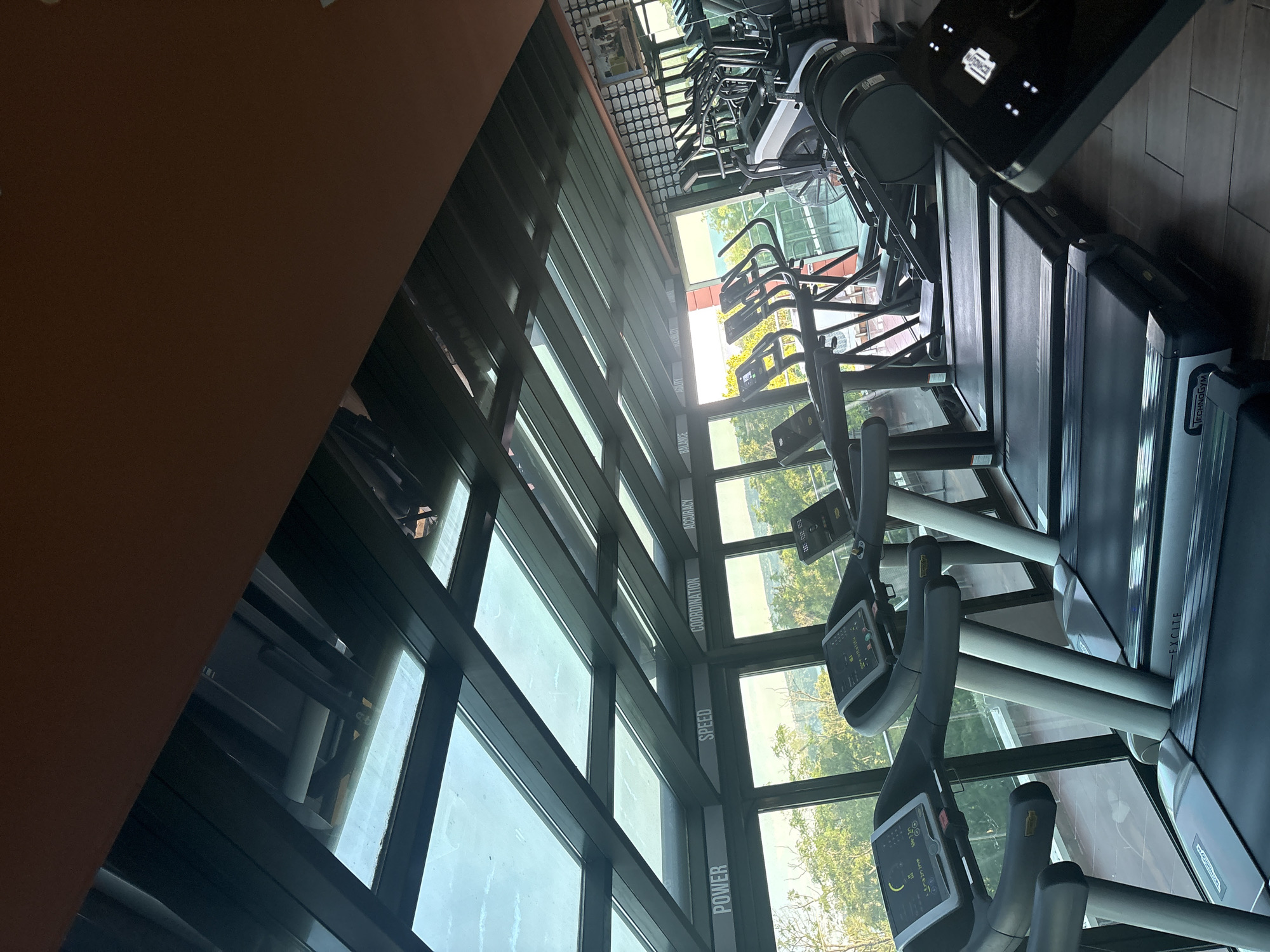 The image depicts an indoor gym with a row of treadmills positioned along a wall of windows. The treadmills are modern and black with grey accents, featuring digital consoles. Some of the consoles display workout metrics. Beyond the windows, lush green trees and a distant body of water, possibly the ocean, are visible under a bright sky, suggesting a scenic location. The gym ceiling is made of glass panels supported by dark metal beams, allowing natural light to flood the space. Words like "POWER," "SPEED," "COORDINATION," and "ACCURACY" are printed on the interior walls above the windows. Other exercise equipment, including ellipticals and a fan, are partially visible in the background. The flooring appears to be wood or laminate. No people are present in the image.