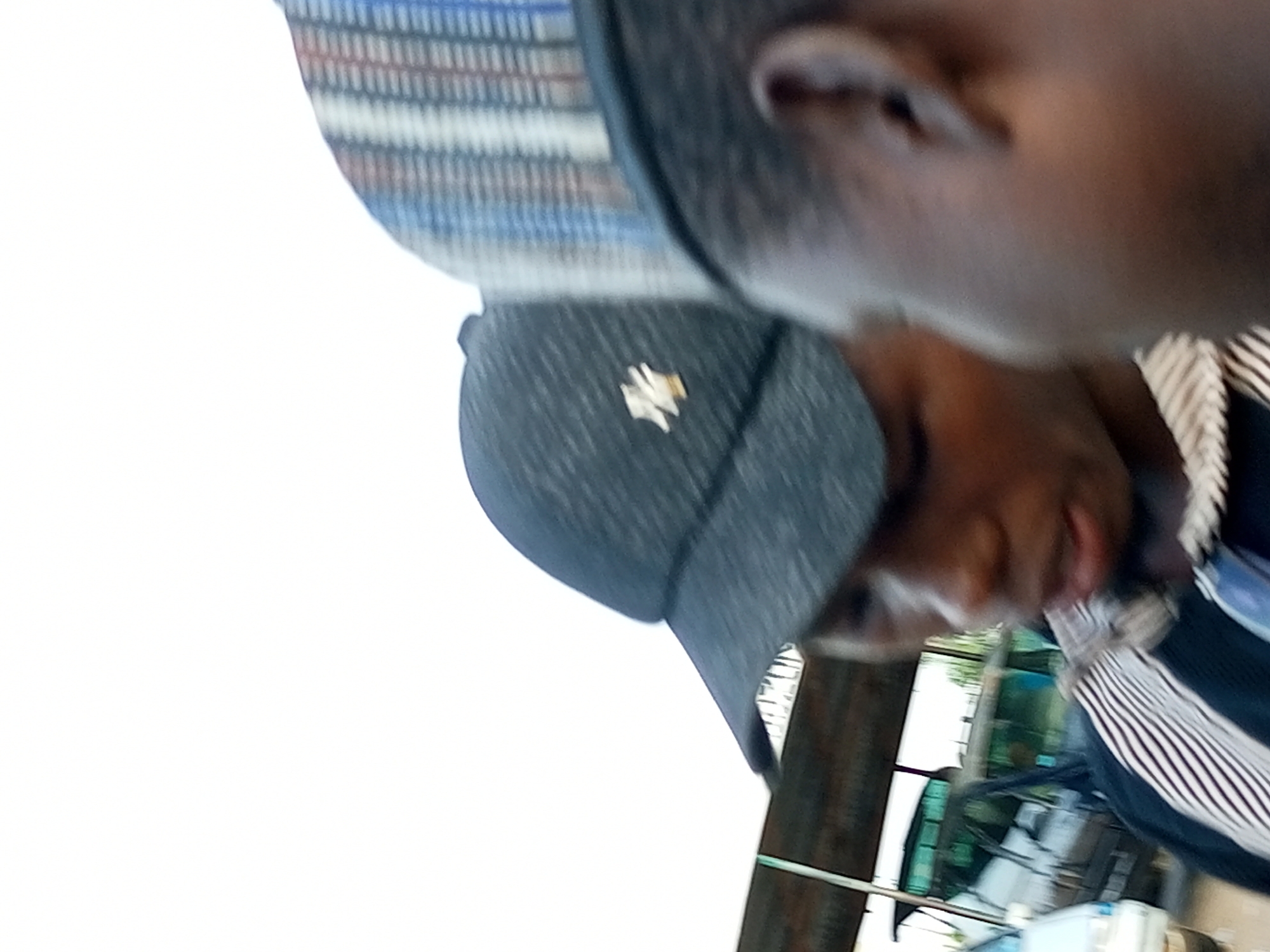 This is a close-up, rotated image of a person wearing a dark baseball cap with a light-colored emblem. The person's face, particularly their ear and part of their cheek, is visible. Behind them, a striped garment and some indistinct background elements can be seen. The image is likely taken outdoors, possibly in the city of Gombe, Nigeria, given the context provided.