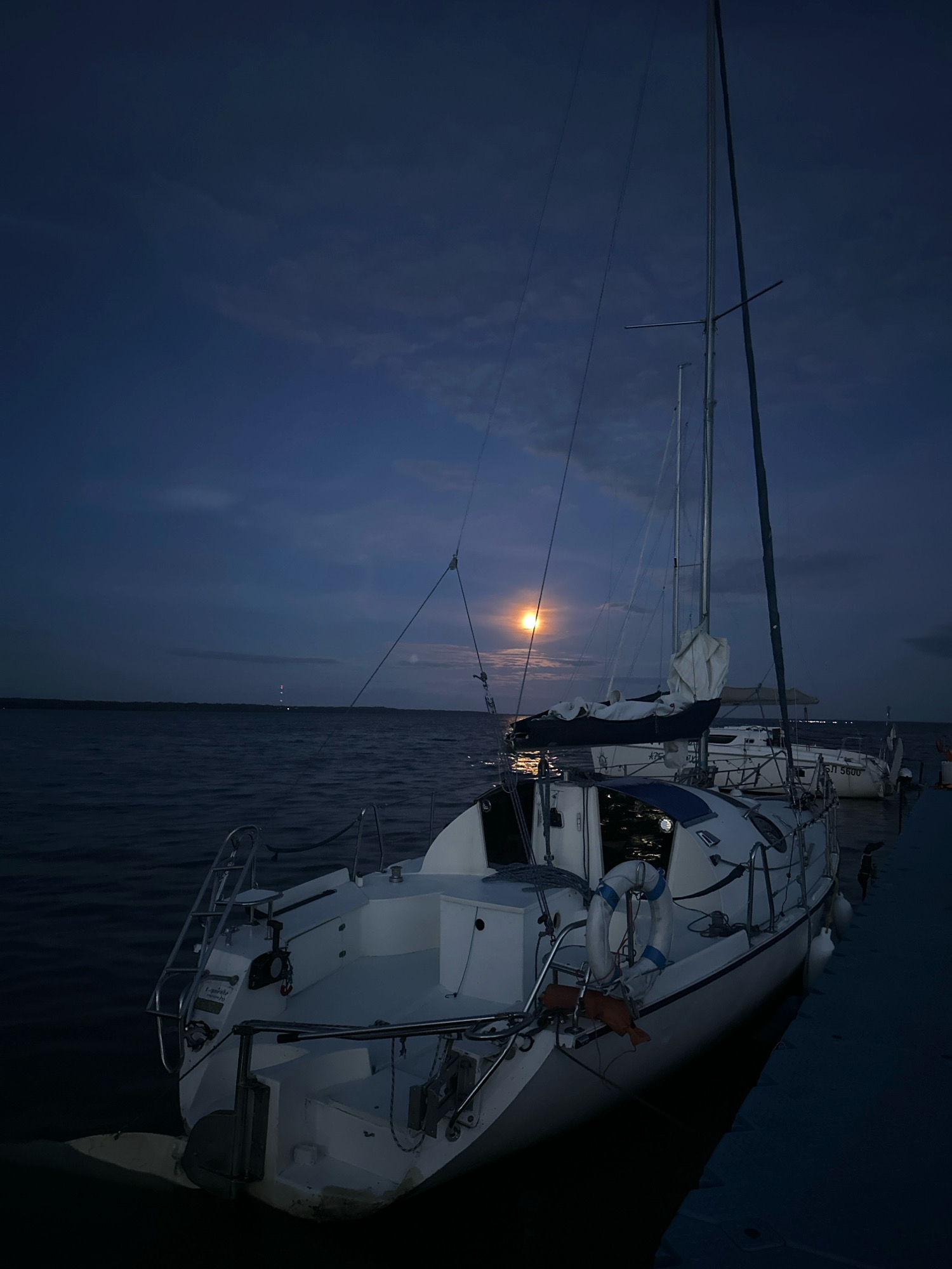 The image depicts a nighttime scene at a marina in the Narač Rural Council, Belarus. A white sailboat is docked on the right side of the frame, with its mast reaching towards a dark blue sky. The full moon is visible in the sky, casting a soft glow on the water and the boat. The water is calm and reflects the moonlight. In the distance, a faint light can be seen, possibly from a lighthouse or another boat. The scene is serene and peaceful.