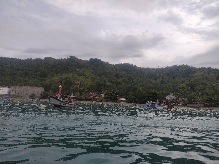 The image presents a tranquil coastal scene in Watulimo, Indonesia, under an overcast sky. The foreground is dominated by calm, deep greenish-blue water with gentle ripples.
Several boats populate the water and shoreline. In the mid-ground, a traditional Indonesian fishing boat, likely a *perahu*, is prominently anchored, tilting slightly to one side. Its hull is light blue and white with red trim on its tall mast structure. Faintly visible text, possibly "KS7" or similar, can be seen on its side. Closer to the sandy shore, numerous smaller fishing boats, some featuring colorful accents, are either moored in the shallow waters or pulled onto the beach.
The background features a verdant, densely forested hill rising steeply from the coast, covered in lush tropical vegetation. Along the base of this hill, a small settlement is visible. On the left, a light-colored, multi-story building with a dark roof stands next to a large, weathered concrete structure. Further to the right, smaller houses with reddish-brown roofs are nestled among the trees along the waterfront.
The sky is entirely overcast with soft grey clouds, suggesting a cloudy day and diffusing the light evenly across the landscape. No people are clearly visible in the scene. The overall impression is one of a quiet, working fishing community integrated with its natural surroundings.
