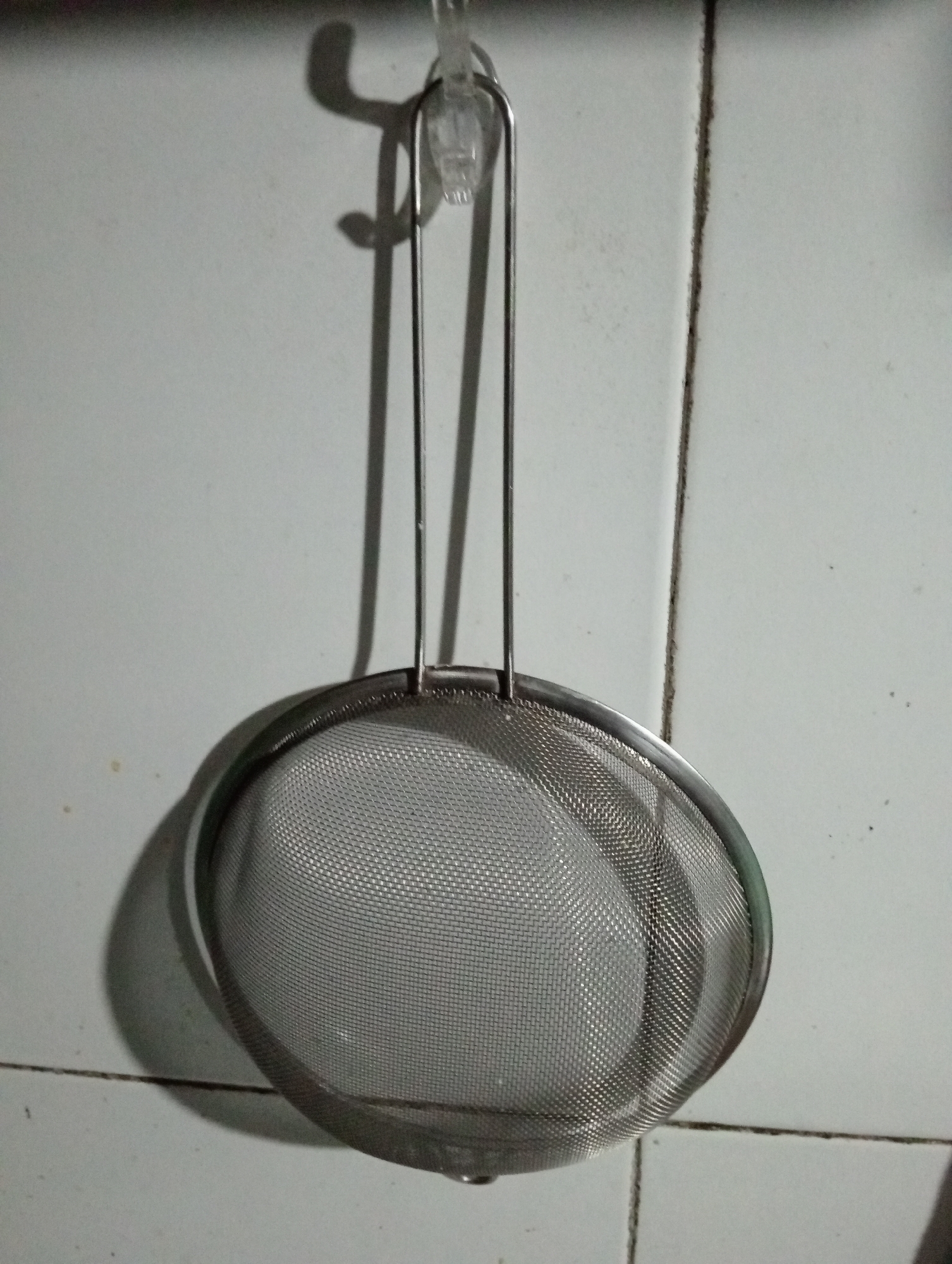 A metal mesh sieve hangs from a clear plastic hook attached to a tiled wall. The sieve has a round basket and a long handle. The wall tiles are white and appear to be in a kitchen or similar area. Shadows are cast on the wall from the sieve and hook. The image depicts a common kitchen utensil hanging in what appears to be a domestic setting in Trenggalek, Indonesia.