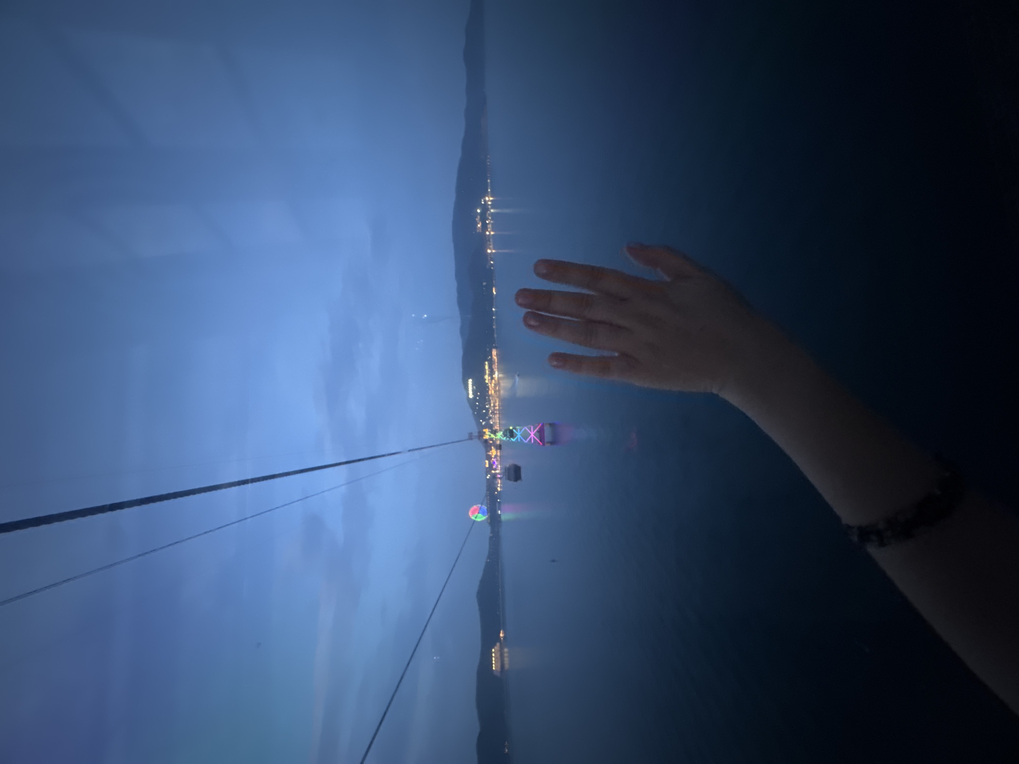 The image is a first-person perspective from inside a cable car, looking out over the water at dusk in Nha Trang, Vietnam. The sky is a deep blue, with some clouds. The water is calm and dark, reflecting the lights of the city. In the distance, there are several illuminated structures, including a Ferris wheel and what appear to be buildings and boats along the coastline. A hand is pressed against the window on the right side of the frame, suggesting the viewer is looking out from within the cable car. The cable car wires are visible diagonally across the top of the image. The overall impression is one of a serene and illuminated evening view of the coastal city.