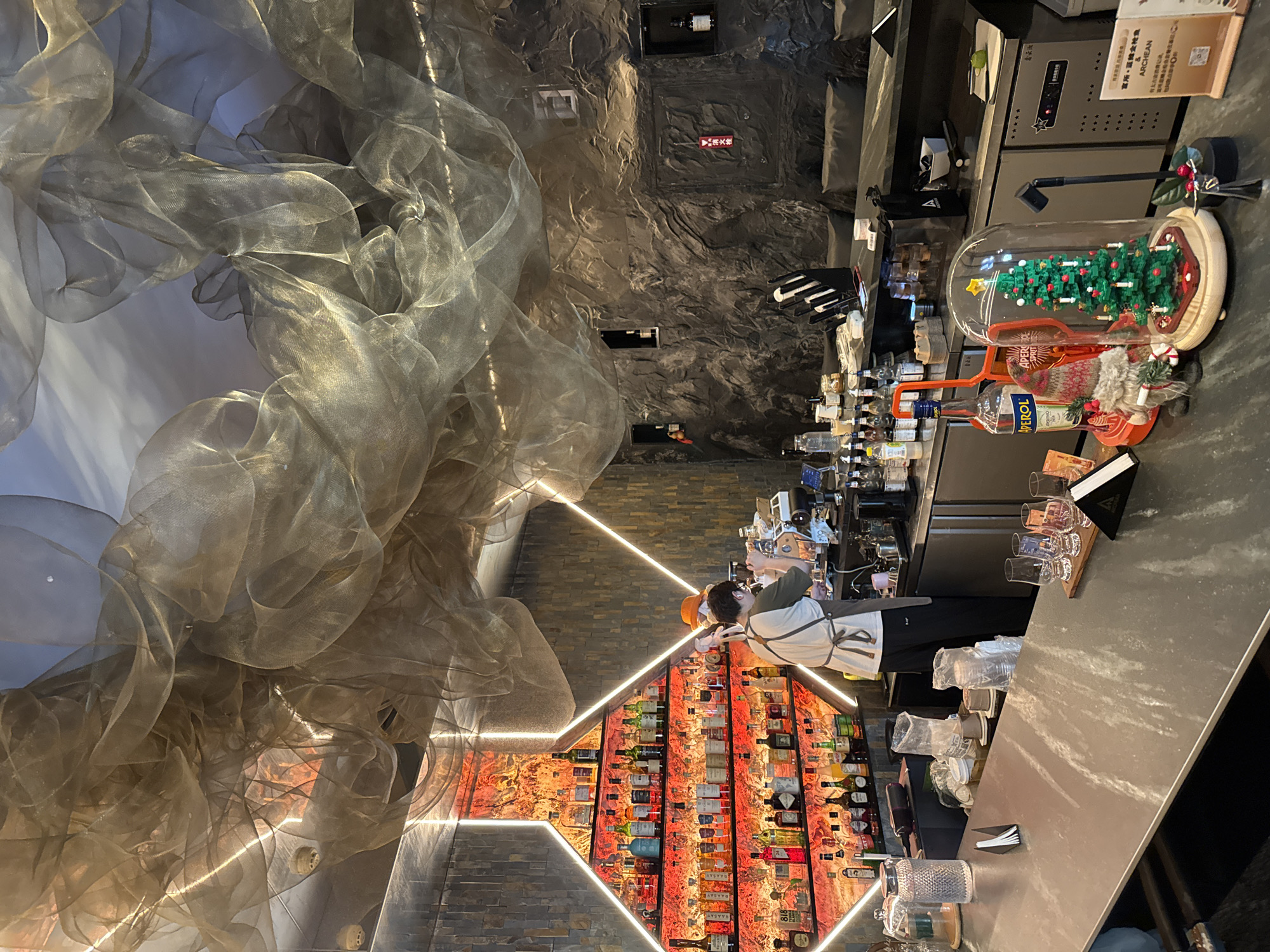 A bartender stands at a bar that serves a variety of alcoholic beverages. Behind the bartender is a wall containing numerous bottles of alcohol. The wall has a red/orange glow and is encased in bright white lines that make a diamond shape. Above the bartender is a rock-like ceiling. To the left of the bar is a cloud-like sculpture that floats down from the ceiling, and the photo was taken in the Jinjiang District, China.