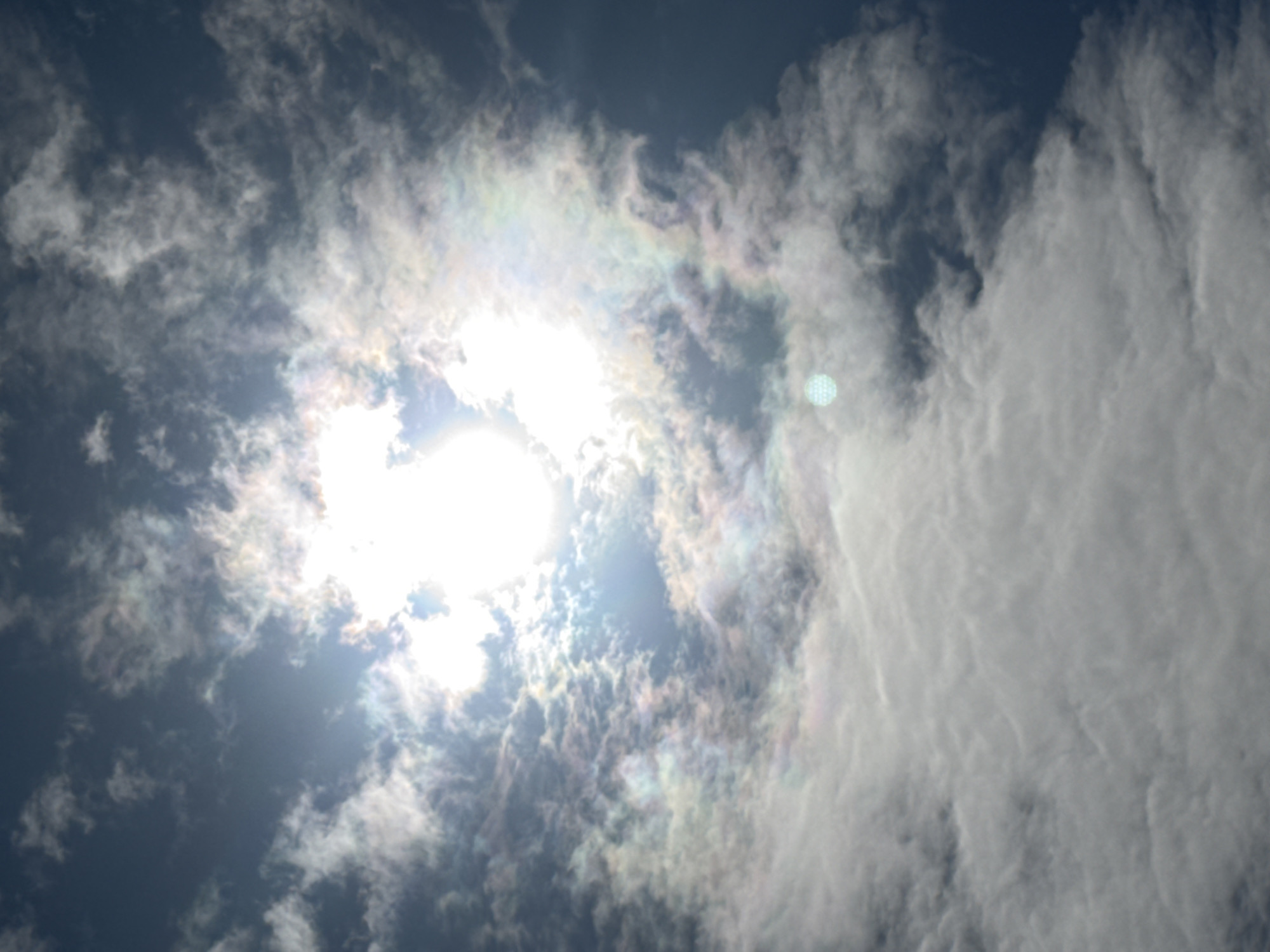 The image shows a sky with clouds and the sun. The sun is partially obscured by clouds, creating a bright halo effect with iridescent colors. The clouds are varied in texture and density, with some appearing wispy and translucent while others are thick and opaque. The sky transitions from a deep blue in the upper right corner to lighter tones where the sun is present. The overall impression is of a dynamic sky, possibly at midday, with the sunlight interacting with atmospheric particles to create optical phenomena. The image does not contain any elements that suggest a specific location within the city of Putiniškės, Lithuania, or any human presence.