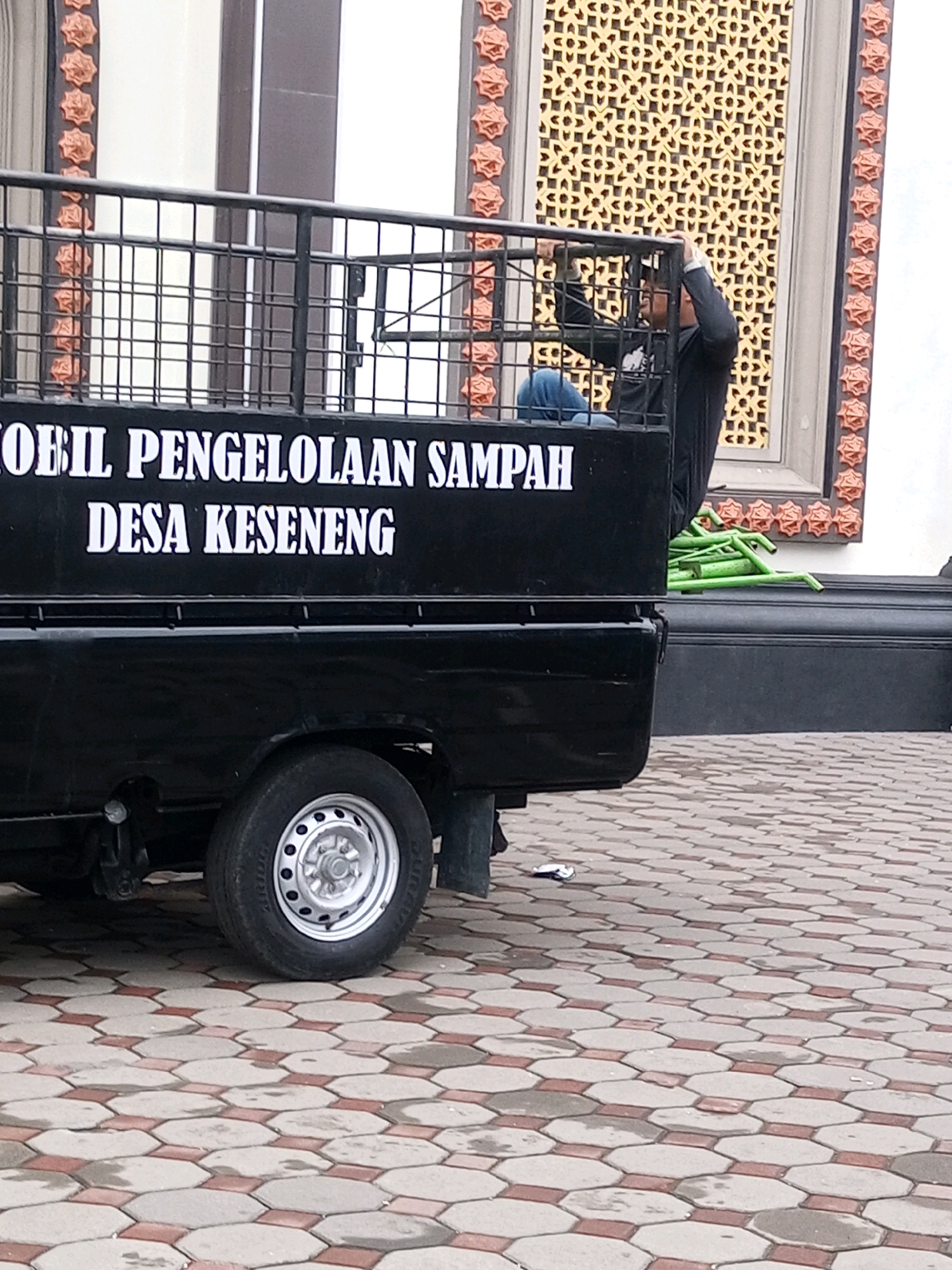 A black pickup truck with a cage on the back is parked on a hexagonal paved walkway. The words "MOBIL PENGELOLAAN SAMPAH DESA KESENENG" are printed in white on the side of the truck, indicating it is a waste management vehicle for the village of Keseneng. A person in a black long-sleeved shirt and blue jeans is partially visible inside the cage, reaching up with one hand. Several green tubes or poles are piled to the right of the truck. The background features a building with decorative panels and columns.