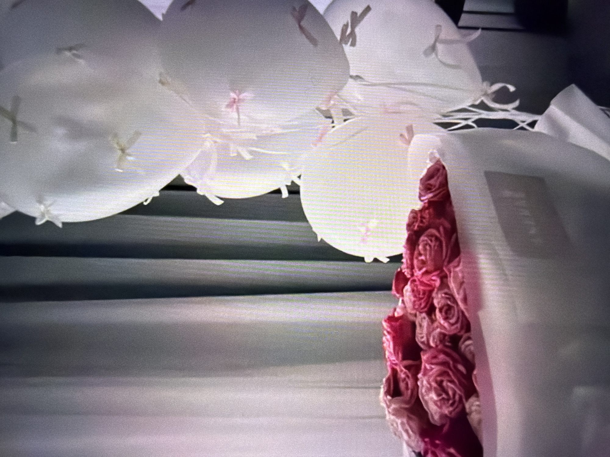 The image shows a close-up, low-angle view of a bouquet of pink roses partially wrapped in white paper. The roses are tightly packed and have a deep pink to lighter pink hue, with visible texture and detail in their petals. The white wrapping paper is semi-sheer, allowing some of the bouquet's color to show through, and is tied with a light-colored ribbon.
In the background, there are several white balloons, some with small pink or light-colored ribbons attached. The balloons appear to be hanging or clustered together. The background also features horizontal lines, suggestive of blinds or a textured surface, in muted gray tones. The overall lighting is soft and diffused, with some shadows. The scene appears to be indoors, possibly set up for a celebration or event. There is no discernible time of day or weather information. No people are visible. No text is clearly legible in the image.