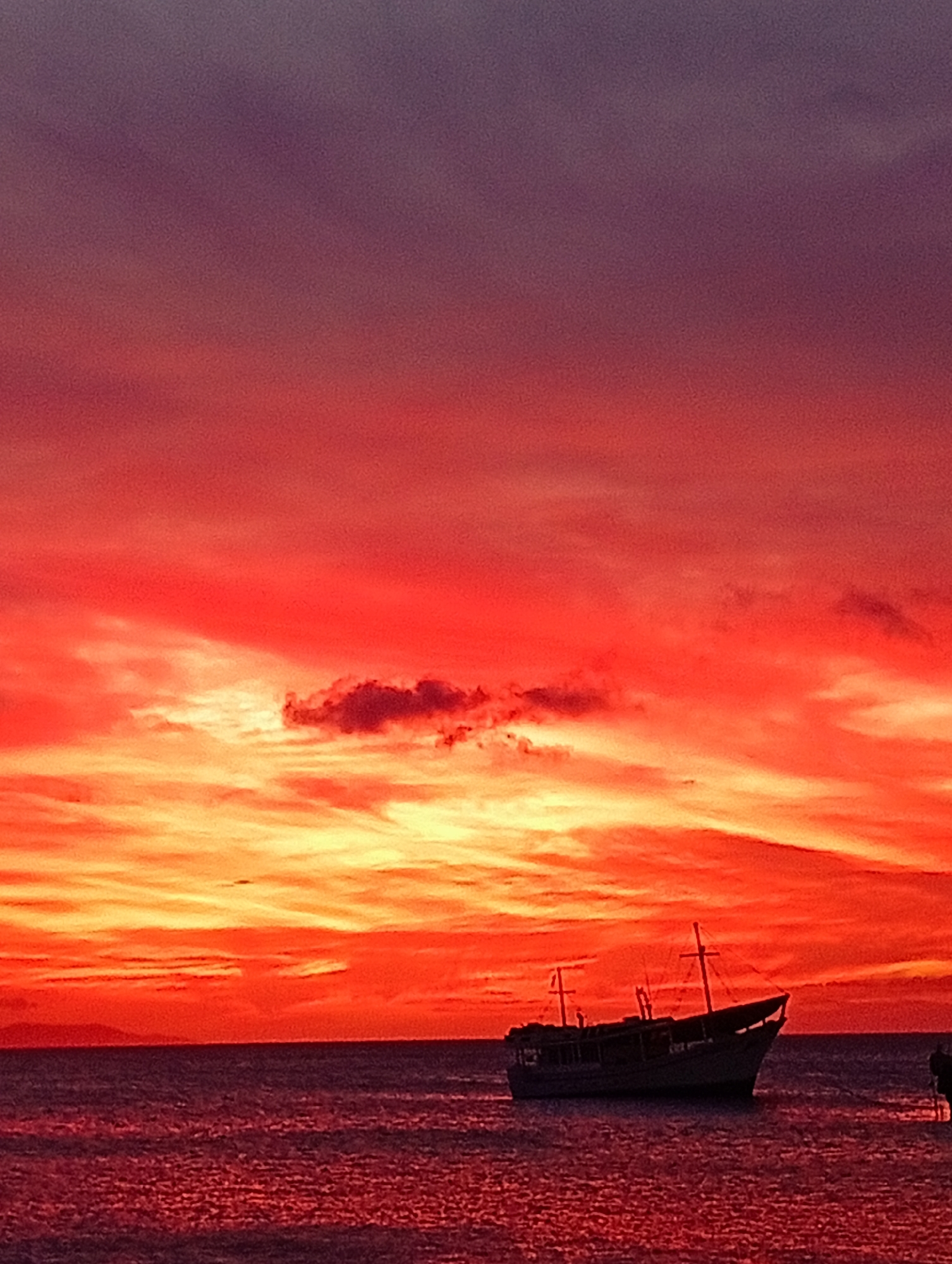 The image displays a coastal sunset scene over the Caribbean Sea in Juan Griego, Venezuela. The sky occupies approximately two-thirds of the frame, exhibiting a gradient of colors from deep purple at the top, transitioning through vibrant red and orange, to bright yellow-orange at the horizon. Scattered clouds are present, rendered in dark silhouettes or illuminated with red and orange hues. The lower third of the image shows the ocean, reflecting the sky's colors with a dark reddish-purple tint and subtle surface ripples. A single, dark-hulled vessel with visible mast structures, consistent with a fishing boat or small cargo ship, is centrally positioned on the water, rendered in silhouette. A faint, dark landmass is visible on the distant horizon line to the left. No individuals or specific actions are discernible within the frame.