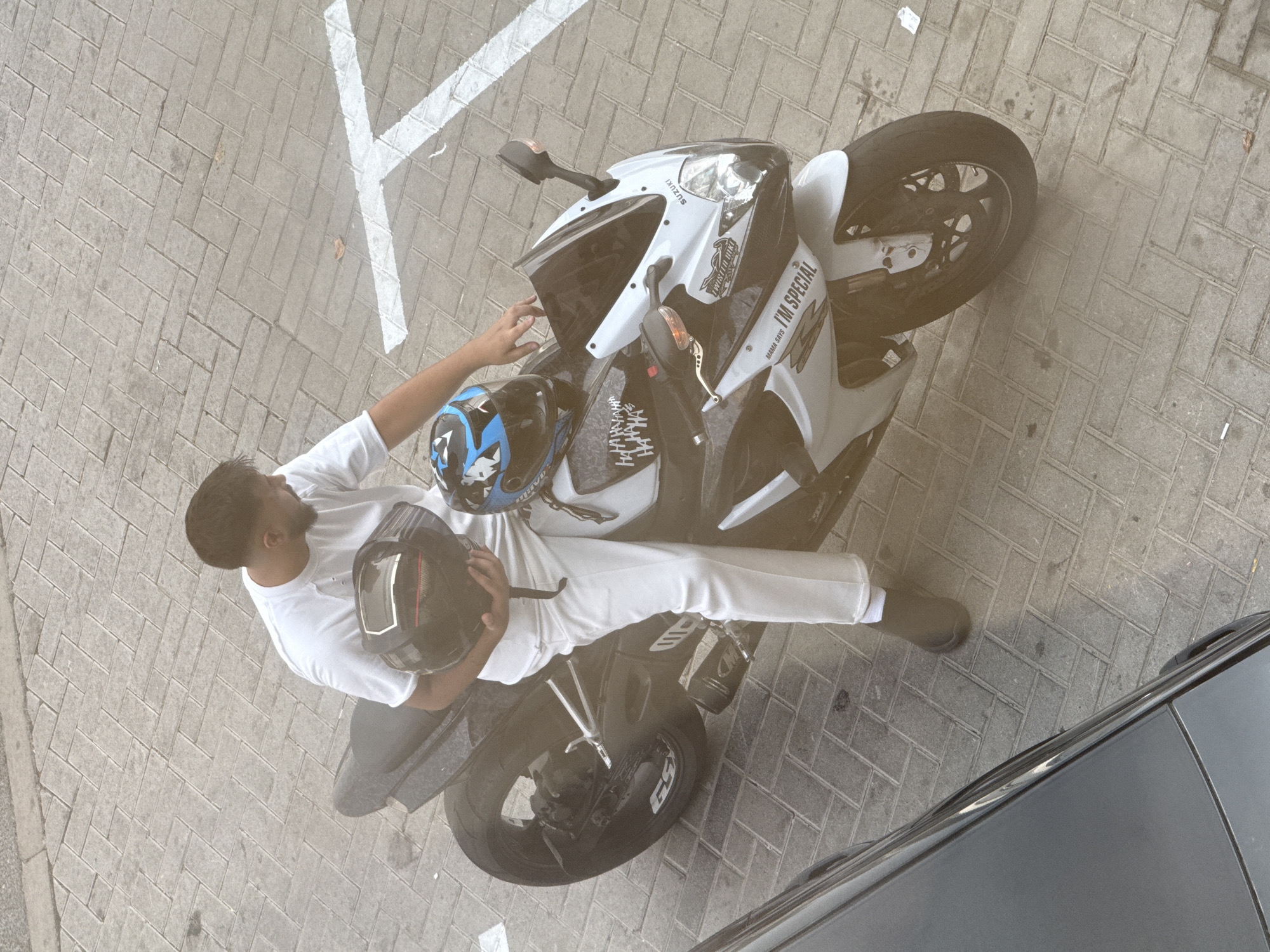 In Dubai, United Arab Emirates, a person is standing next to a white and black Suzuki sportbike parked on a paved surface with a herringbone pattern. The individual, dressed in a white t-shirt and white trousers with black boots, holds a black motorcycle helmet in their right hand and is reaching towards a blue and black helmet positioned on the motorcycle's seat. The sportbike features several decals, including "MAMA SAYS I'M SPECIAL" and "TWISTED LIQUI". The ground is marked with white lines, suggestive of a parking area. A portion of a dark gray vehicle is visible in the lower left corner of the frame.
