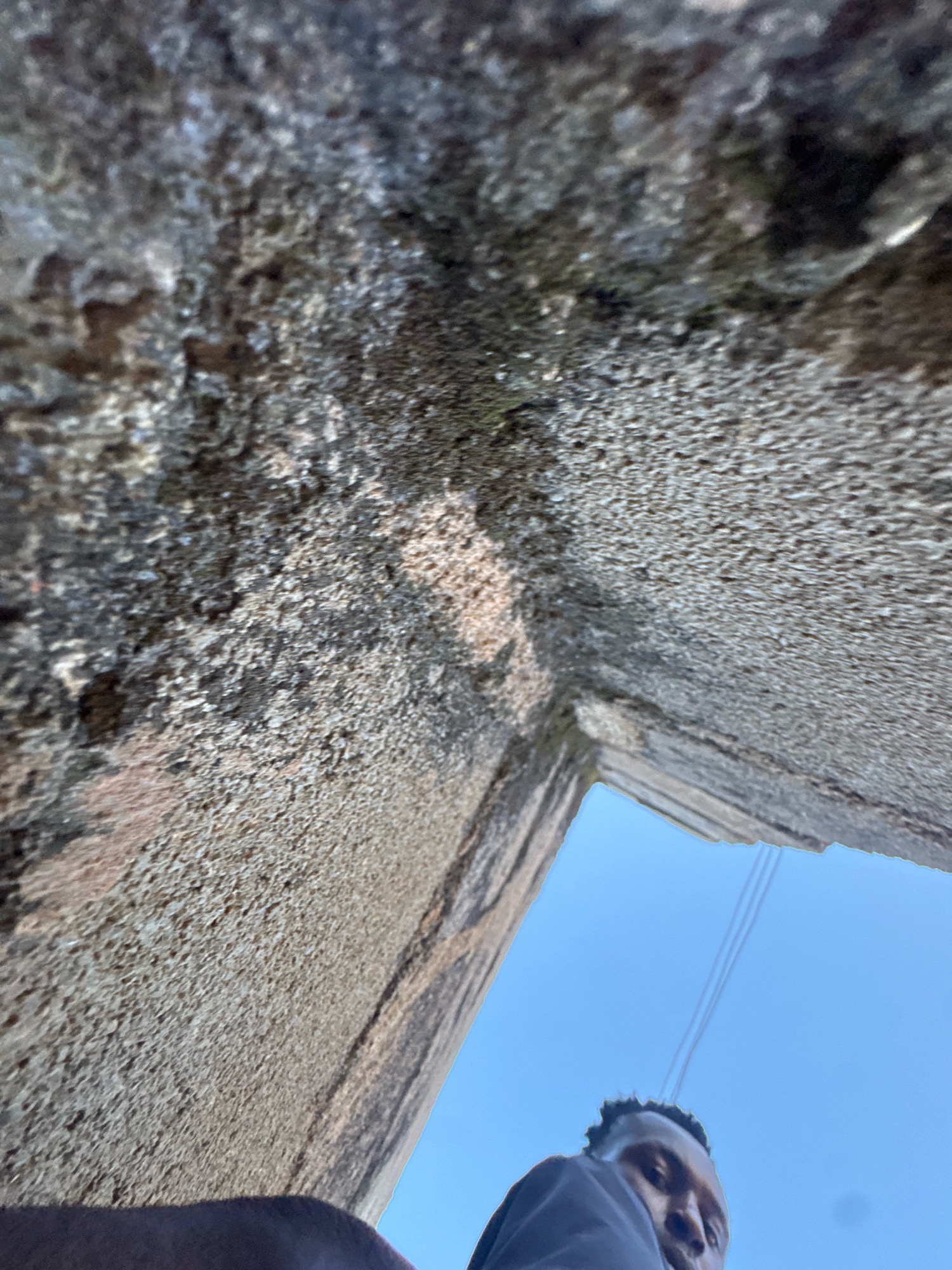 The image is an upward-facing selfie taken from a low angle, looking towards the sky. The majority of the frame is occupied by rough, textured stonework, possibly a wall or the underside of a structure, with a corner visible in the center right. The stonework is predominantly gray and brown with some areas appearing damp or weathered. A bright blue sky is visible through a rectangular opening in the stonework. In the bottom portion of the image, a person's face and shoulder are visible, looking directly at the camera. The person has dark skin and short, dark hair. They are wearing a dark-colored garment. Three faint, dark lines are visible against the sky, suggesting power lines or wires. The overall impression is one of being in an urban environment, possibly looking up from a confined space or under a bridge.