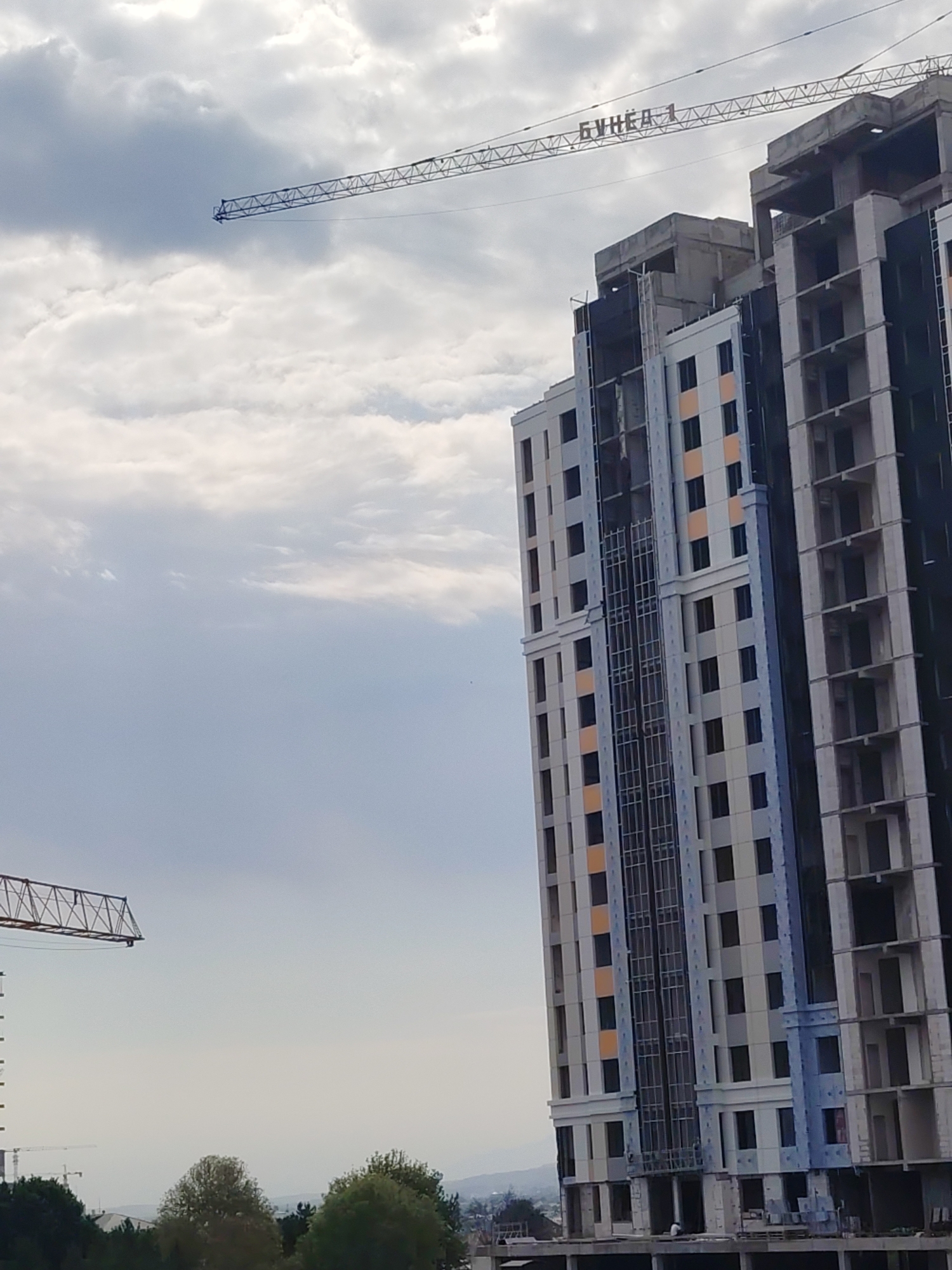The image shows a construction site in Khujand, Tajikistan, featuring a partially built high-rise building and a construction crane. The building is a multi-story structure with a facade partially covered in light-colored panels and some sections revealing exposed concrete framework. Several windows are visible. A large construction crane, with its boom extended over the building, has Cyrillic text that reads "БУНЁД 1" on its jib. The sky is overcast with scattered clouds. In the foreground, trees and other vegetation are visible, along with what appears to be another smaller construction crane in the distance. The overall scene depicts an active construction project in an urban or semi-urban environment.