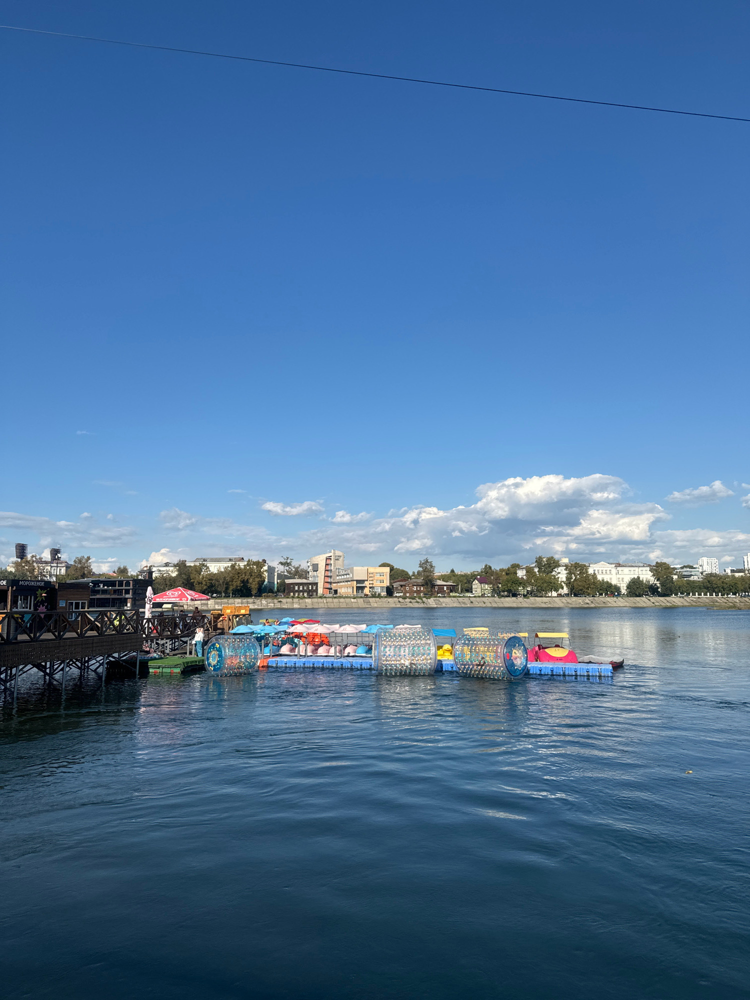 A wide-angle, eye-level view captures a clear blue sky with scattered white clouds above a calm, dark blue body of water. In the foreground, the water surface exhibits gentle ripples. A wooden pier extends from the left side of the frame, featuring a railing and a structure with a sloped roof. Attached to the pier are green floating platforms supporting several colorful inflatable attractions. These include large, transparent cylindrical water rollers, a yellow watercraft with a pink canopy, and a row of orange and pink flotation devices with backrests. A few people are visible on the pier and on the floating platforms, some appearing to be operating the attractions. Across the water, a tree-lined embankment separates the water from a cityscape. The city features a variety of buildings, including multi-story structures with light-colored facades and some smaller, older buildings. The scene depicts a recreational area by the water on a sunny day. The location is identified as the Angara River embankment in Irkutsk, Russia, specifically near the recreation area on the river.
