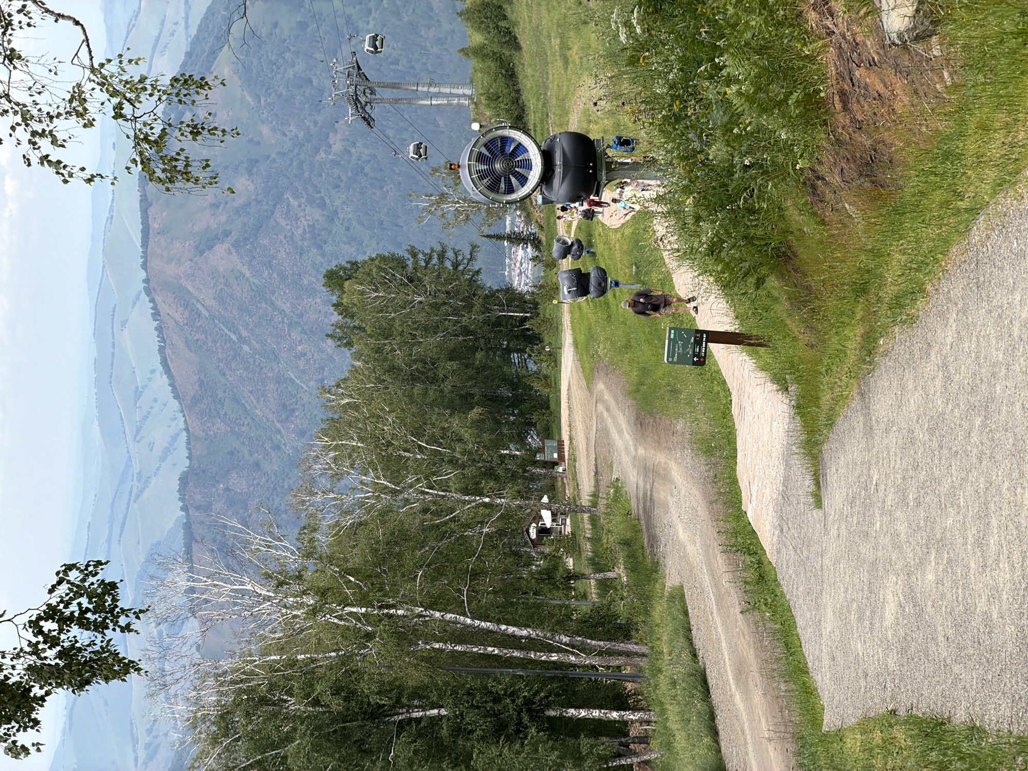 The image depicts a mountain landscape with a winding gravel path in the foreground, leading uphill. To the right of the path, several large snow cannons are visible, with one prominently featured in the center. Several people are walking up the path. In the background, there are dense green trees, a ski lift with gondolas, and rolling hills fading into the distance under a hazy sky. The location is identified as Озерное (майминский), Russia. The scene appears to be a tourist area, possibly a ski resort during the off-season.