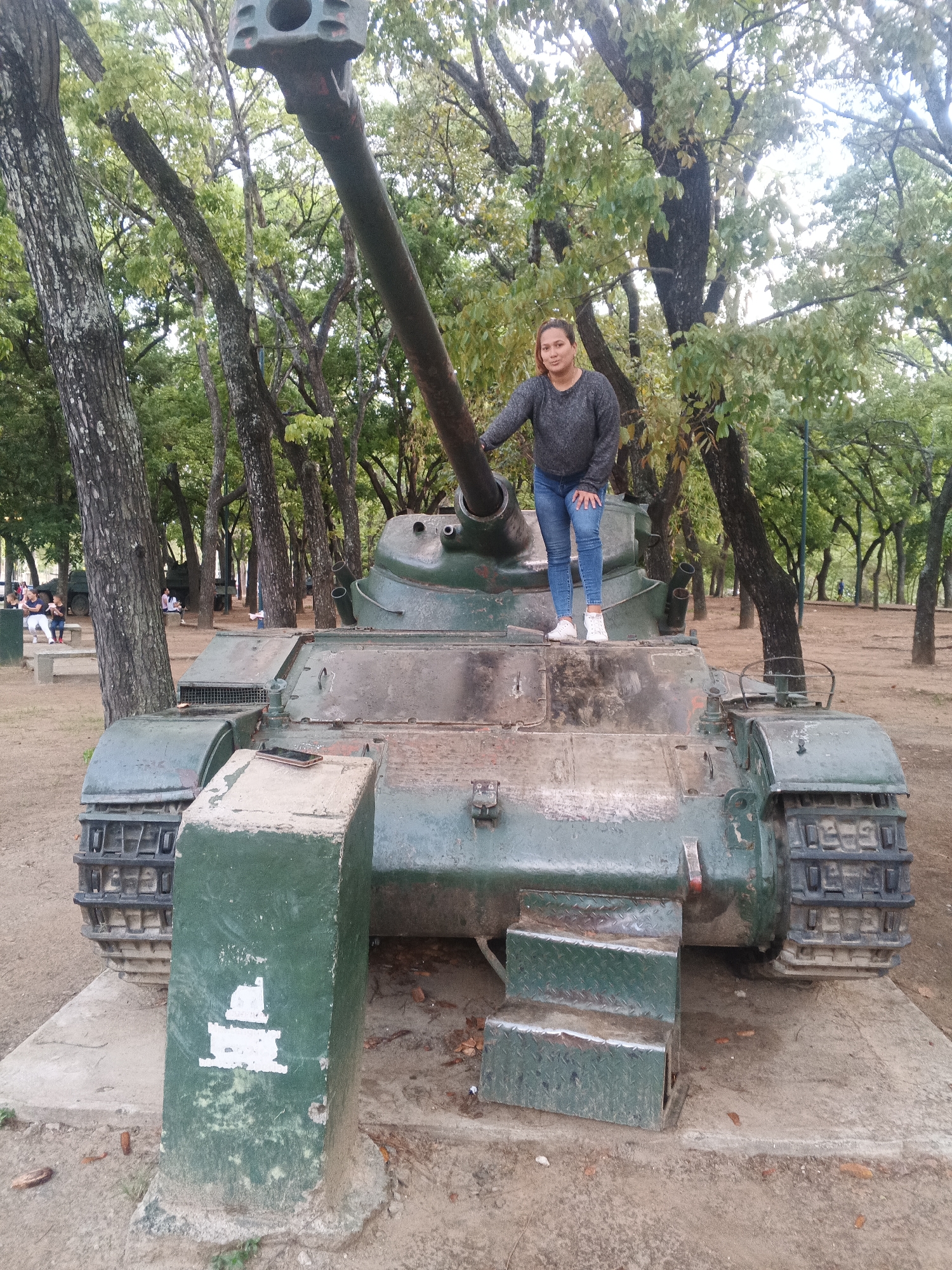 A woman in blue jeans and a gray sweater stands on the turret of a green military tank in a park-like setting. She is facing forward with her left hand resting on the tank's gun barrel. The tank has an extended gun barrel pointing upward and is positioned on a concrete base with attached steps. A green concrete pillar with a white painted design stands to the left of the tank. Trees with green foliage surround the tank, and people are visible sitting on benches in the background. The scene is set in Caracas, Venezuela.