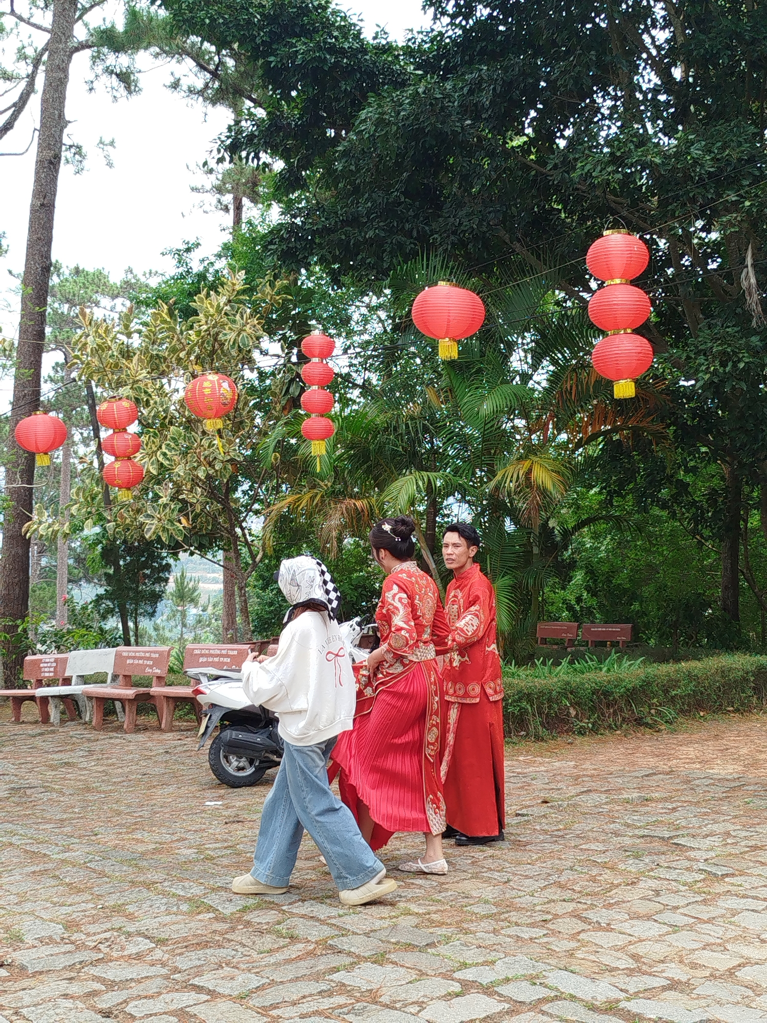 The image depicts an outdoor scene in Da Lat, Vietnam. Three individuals are present: one woman wearing jeans, a white sweatshirt, and a headscarf; another woman in a traditional red dress; and a man in a red outfit, also of traditional style. All three are standing near a scooter. There are red paper lanterns strung in the background. There are park benches. The location is paved with patterned brick.