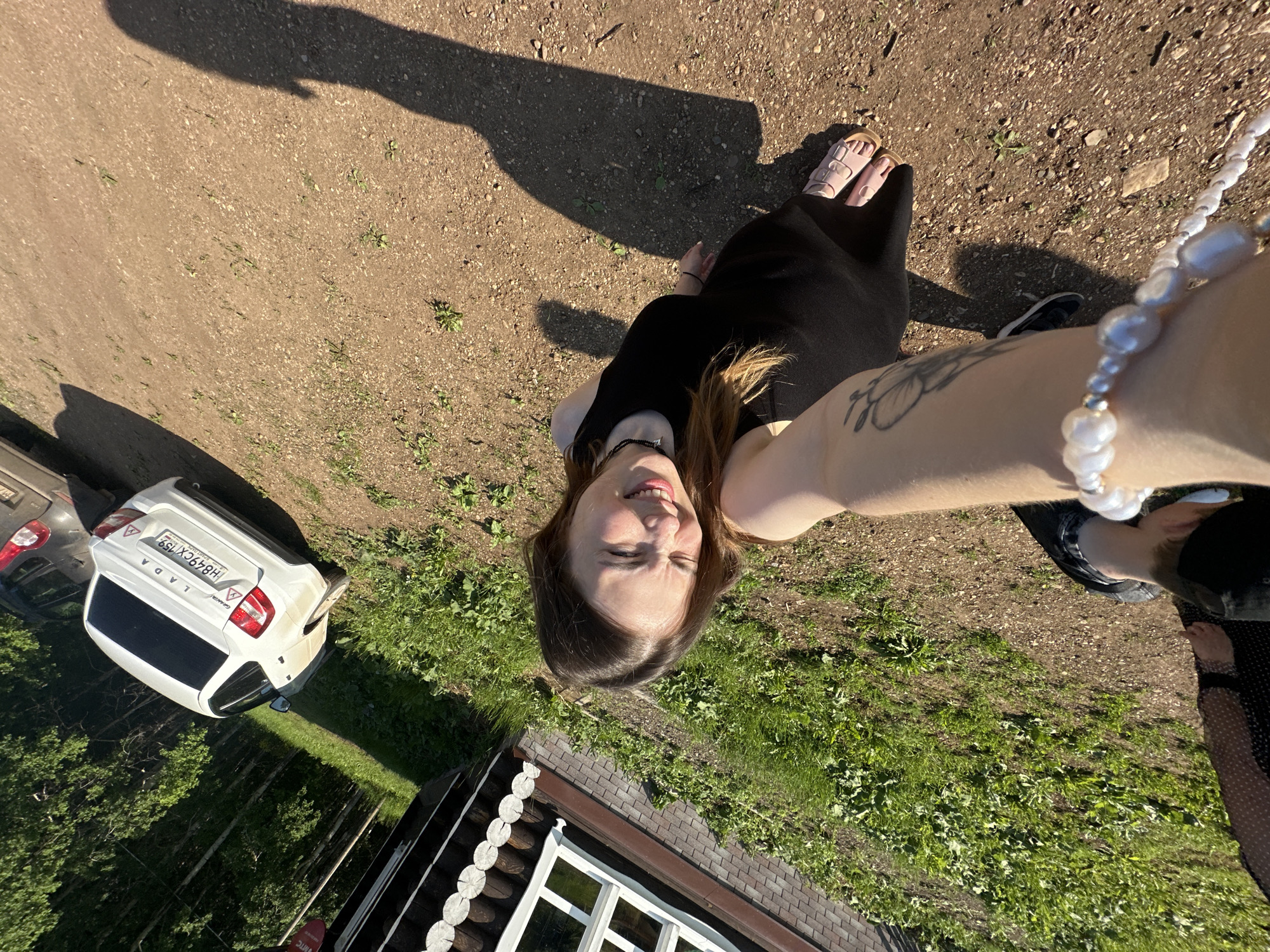 In Bogorodskoe, Russia, a young woman with long brown hair, wearing a black dress and sandals, is captured in a selfie. She is standing on a dirt path with sparse green vegetation. To her left is a wooden house with white-framed windows. A white Lada car is parked on the dirt path behind her. The scene is illuminated by bright sunlight, casting a long shadow of the woman on the ground.