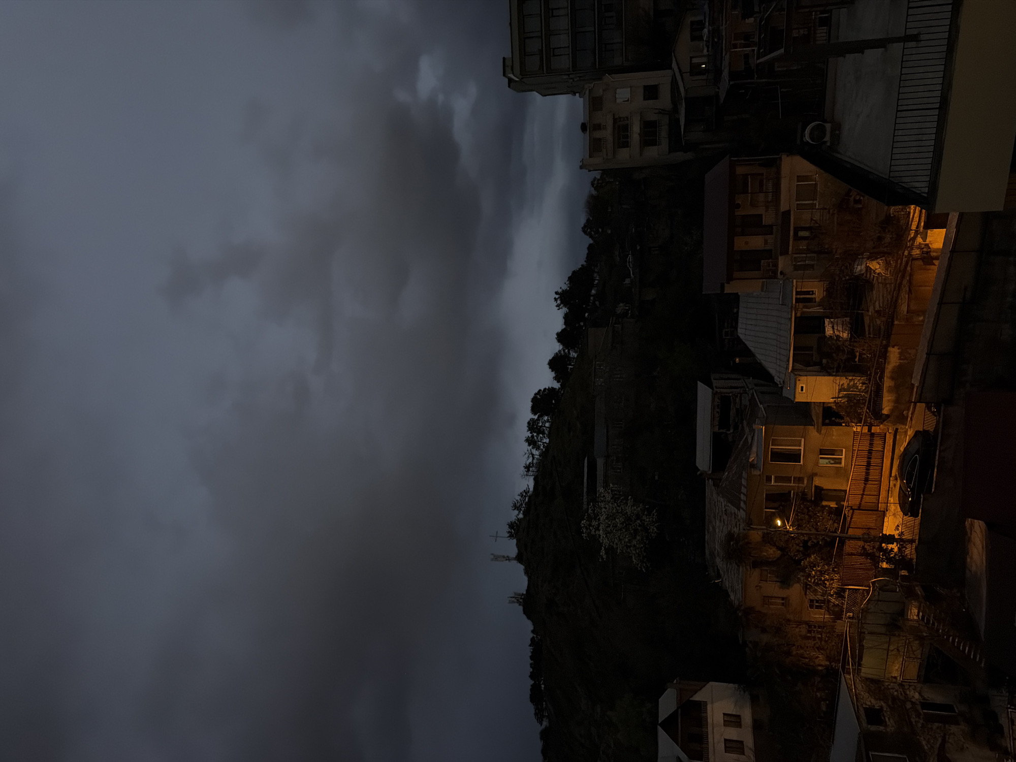 The image depicts a view in Tbilisi, Georgia, likely taken at dusk or night under an overcast sky. The right side of the image shows buildings on a hillside, illuminated by warm artificial light. Several houses are visible, along with a parked car. The left side shows a dark, cloudy sky. A wooded hillside rises in the center of the image. In the upper left, tall buildings are visible.