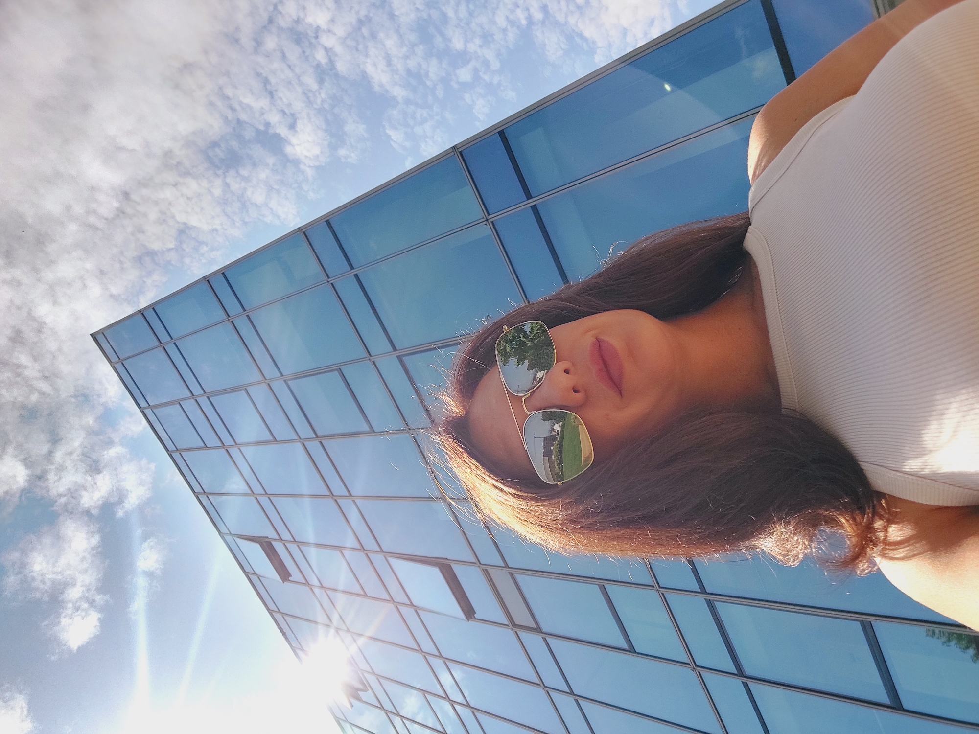A low-angle, medium shot captures a woman from the chest up, looking upwards. She has long, dark brown hair and is wearing aviator sunglasses and a ribbed, light-colored tank top. The sunglasses reflect an outdoor scene with a construction crane and greenery. Behind her, a modern glass-fronted building rises steeply against a bright blue sky with scattered white clouds. The sun is visible on the left side of the frame, casting a lens flare. The image appears to be taken outdoors in an urban environment, possibly in the city of Sofia, Bulgaria.