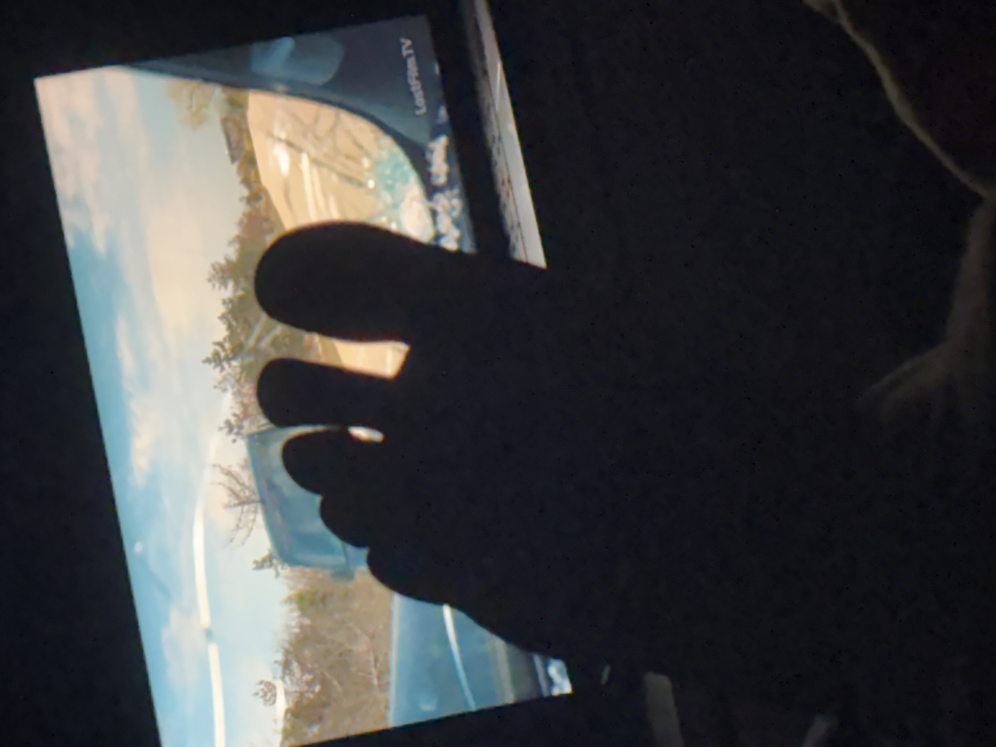 A dark and grainy image shows a screen displaying a first-person perspective view from inside a vehicle driving along a coastal road. A person's toes are in the foreground, partially obscuring the screen. The vegetation on the right side of the road appears sparse, with trees and shrubs typical of a Mediterranean climate. The sky is visible, suggesting it's daytime. There are no identifiable people or interactions except for the toe obscuring the screen. The screen displays "LostFilm TV". The location is impossible to determine without additional context.