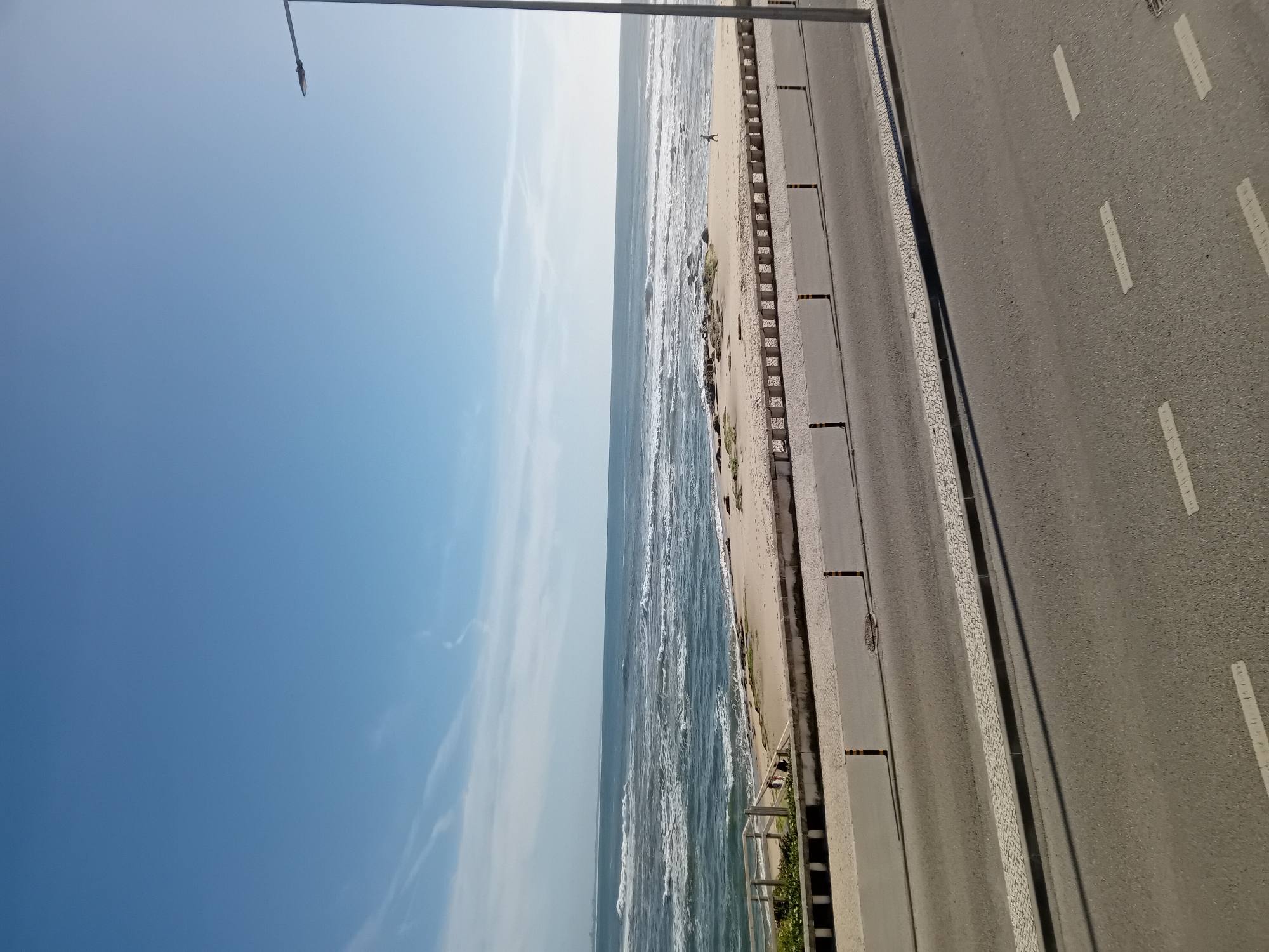 The photograph captures a scene alongside a beach, adjacent to a road. The sky is clear and blue, transitioning to the horizon where it meets the sea. The sea is turbulent, with visible waves. A beach stretches between the sea and a paved road. A person is visible on the beach near the water's edge. The road has white lane markings. A lamppost is located on the left side of the image. The location is identified as Unknown, Unknown.