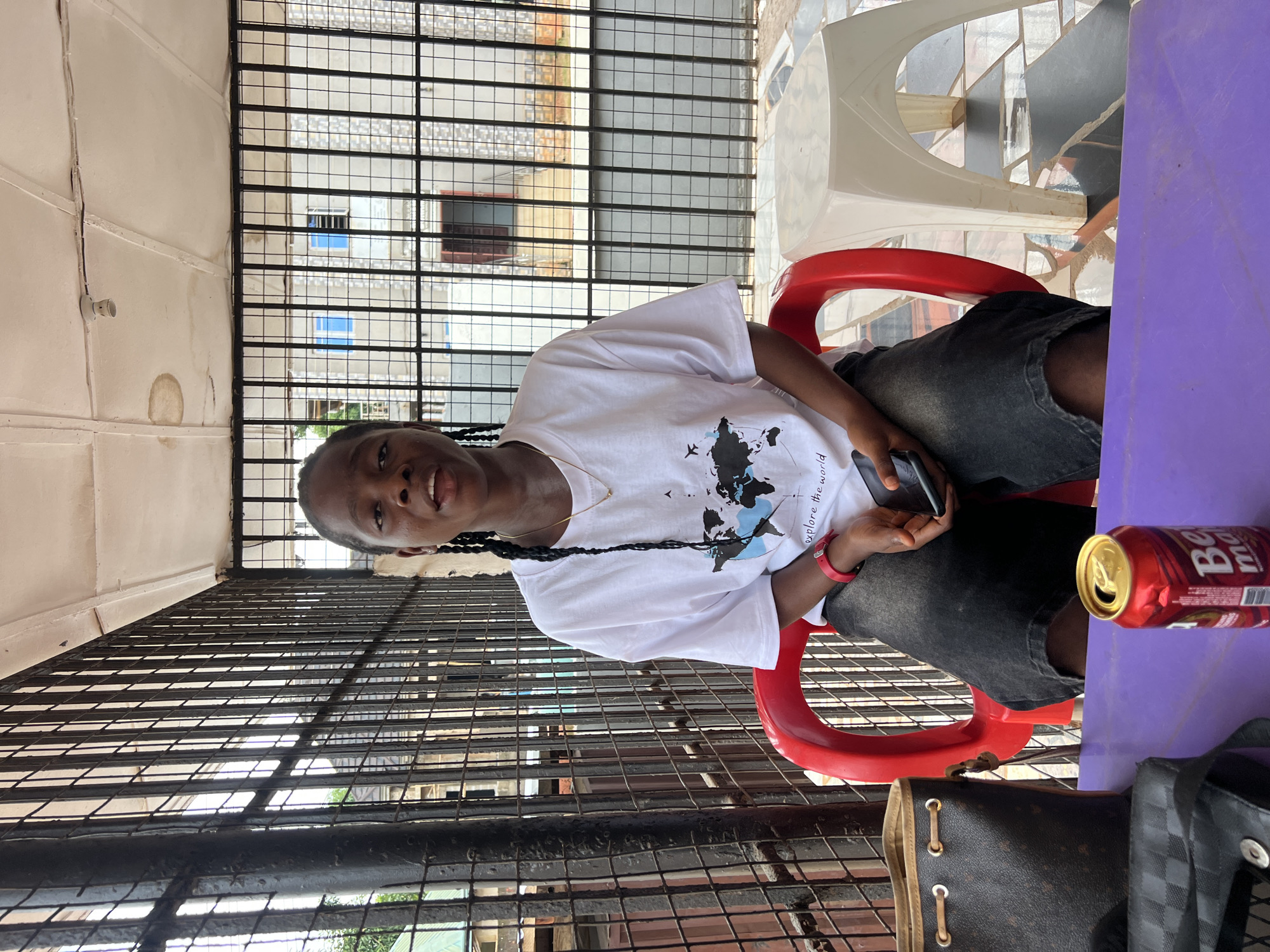 A medium shot captures a young Black woman with braided hair sitting on a red plastic chair in an outdoor covered area, possibly a veranda or patio, in Nkpor, Nigeria. She is wearing a white t-shirt with a black and blue world map graphic and dark denim shorts. A red wristwatch is visible on her left wrist, and she is holding a black smartphone in her right hand. In front of her, on a purple table, sits an open can of "Beer" with a label that is partially obscured. To the left of the can is a brown handbag with gold accents and a black checkered bag. A white plastic stool is positioned to the right of the red chair. The background shows a black metal fence or gate, with a light-colored building and some greenery visible beyond it. The floor is tiled with a checkered pattern. The lighting suggests it is daytime.