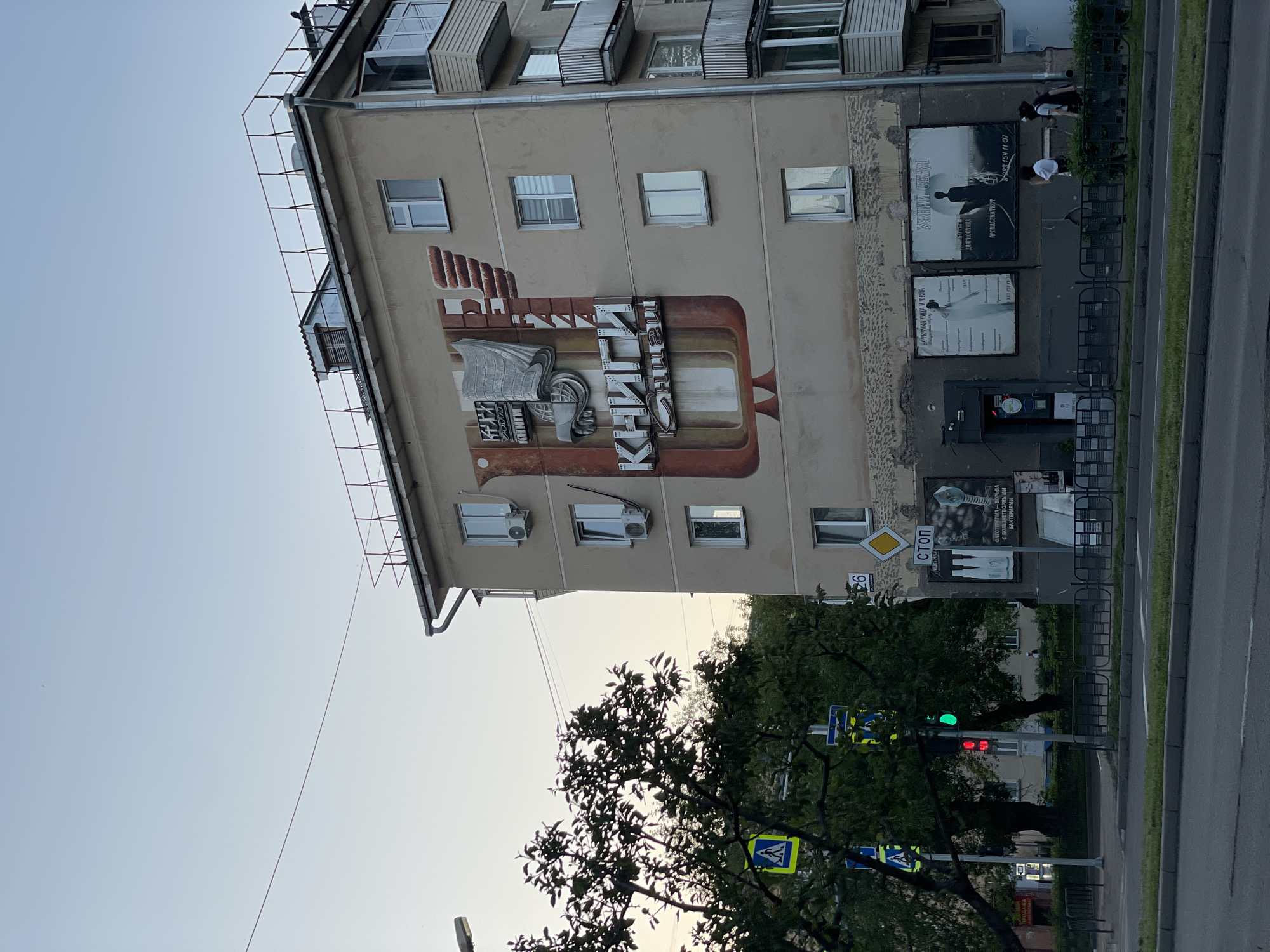 This media file captures an urban street scene in Zheleznogorsk, Russia, likely during the late afternoon or early evening given the soft light and clear sky. The main subject is a multi-story, beige apartment building on the left, featuring numerous windows and several air conditioning units.
A prominent mural decorates the side of the building, depicting stylized books and the word "КНИГИ" (Books) in large Cyrillic script. Below this mural, on the ground floor, are several advertising posters. One poster displays the text "УЗНАЙ СЕБЯ" (Know Yourself), "ДИАГНОСТИКА" (Diagnostics), and "АРОМАКОРРЕКТОР" (Aromacorrection), along with a phone number (8983 154 11 07). Another poster features text about "ЭСТЕТИКА ЛИЦА И ТЕЛА" (Face and Body Aesthetics) and a similar phone number (983 154 0). A third poster mentions "ФАГОТЕРАПИЯ - БОРЬБА С БОЛЕЗНЕТВОРНЫМИ БАКТЕРИЯМИ" (Phage therapy - Fight against pathogenic bacteria). Two people are visible on the sidewalk near these posters, one in a white t-shirt and another in darker clothing.
To the right of the building, a sidewalk borders a street. A black metal fence runs along the sidewalk. Lush green trees partially obscure the view of the street, but a traffic light is visible, displaying a green light for pedestrians and a red light for vehicles. A "СТОП" (STOP) sign is present, along with a yellow diamond-shaped sign and a blue pedestrian crossing sign. The number "26" is also visible on a yellow sign. The road itself is gray asphalt with a white line marking a lane. The sky above is clear and light blue.