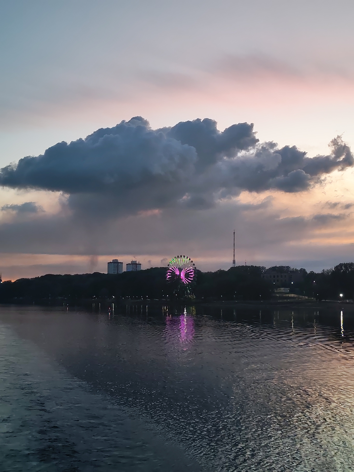 The photograph depicts a body of water reflecting the sky at dusk in Cherepovets, Russia. A large, dark cloud dominates the upper portion of the image, with light pink and orange hues visible in the sky above. A Ferris wheel illuminated with pink and green lights stands on the shore opposite the viewer. The shore is lined with trees and buildings, including a tall radio tower and two high-rise buildings. Lights are reflected in the water. The water's surface is textured with ripples.