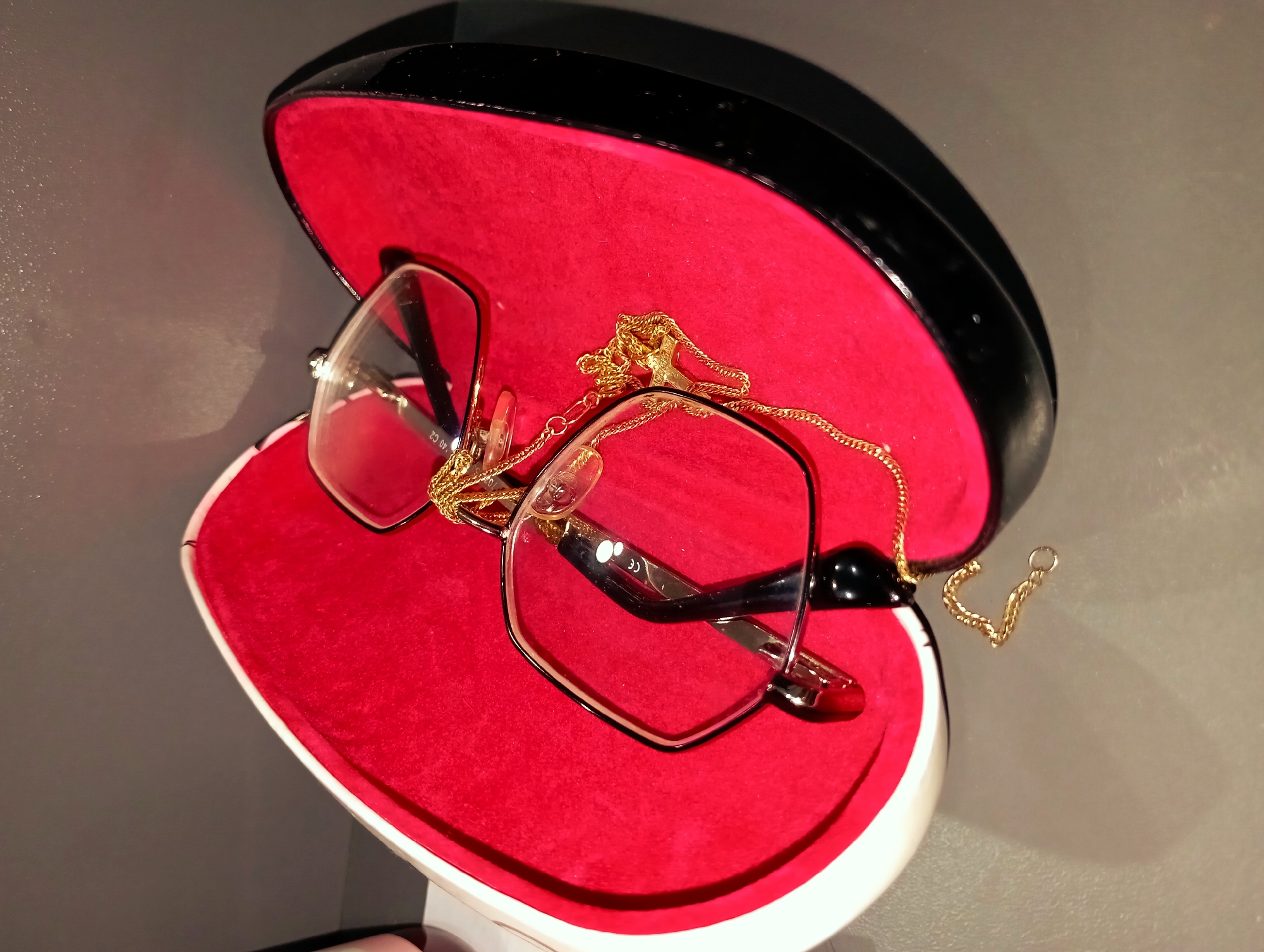 A pair of gold-rimmed eyeglasses lies inside an open eyeglasses case lined with red felt. A gold chain is draped over the glasses and extends outside the case. The case has a black exterior and a white trim. The setting appears to be indoors on a flat surface with a grey background. The location is unknown.