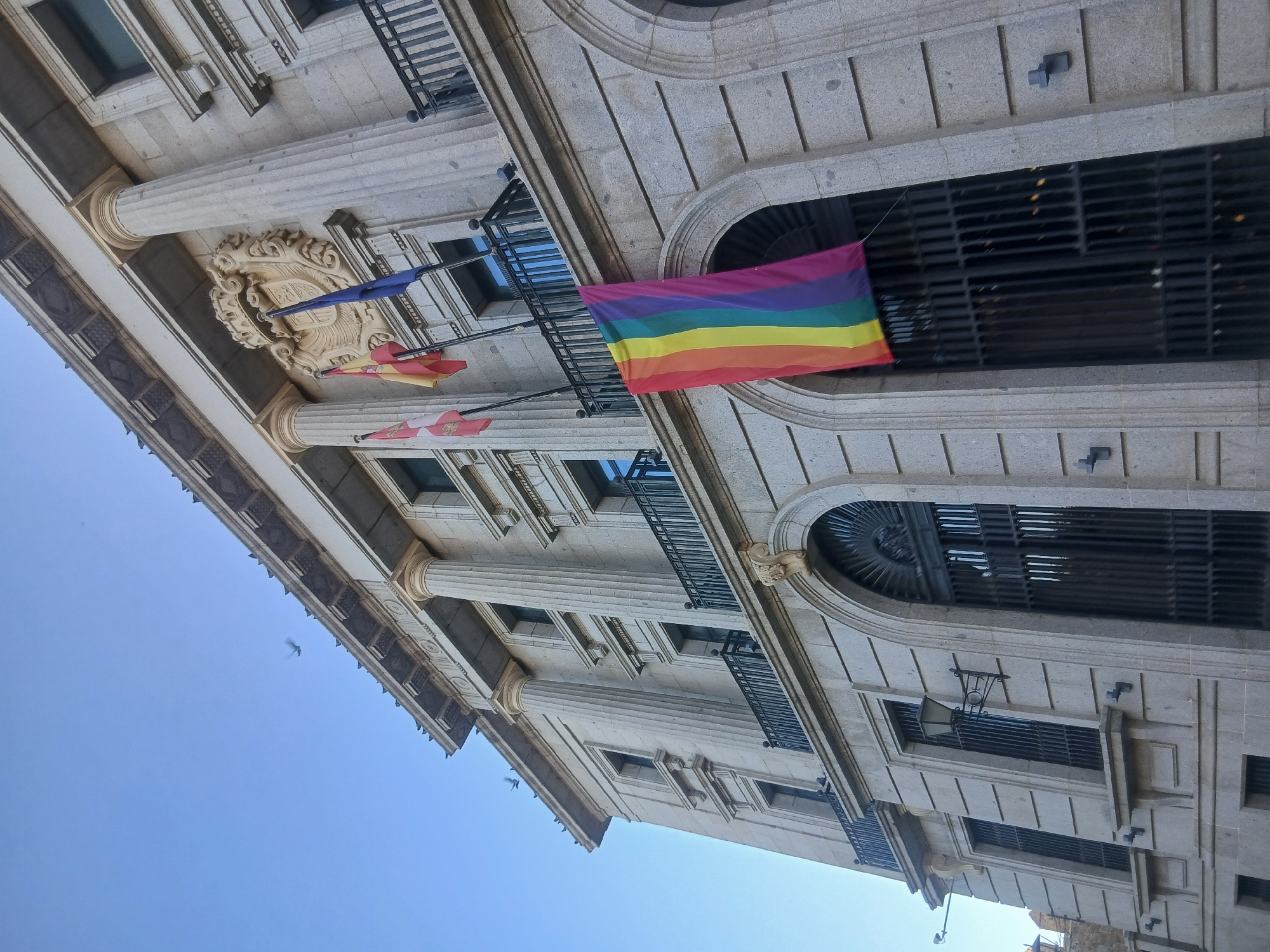 The image shows the facade of a stone building in Ávila, Spain, with a rainbow flag hanging from a balcony. The building is ornate, with classical columns, arched windows, and decorative friezes. Three flags are flying from poles above the balcony: the flag of Spain, another flag with red and yellow stripes, and a blue flag. The rainbow flag, a symbol of LGBTQ+ pride, is prominently displayed below the balcony, partially obscuring a large, dark metal gate. The sky is clear and blue, with a few birds in flight. The perspective is from a low angle, looking up at the building.