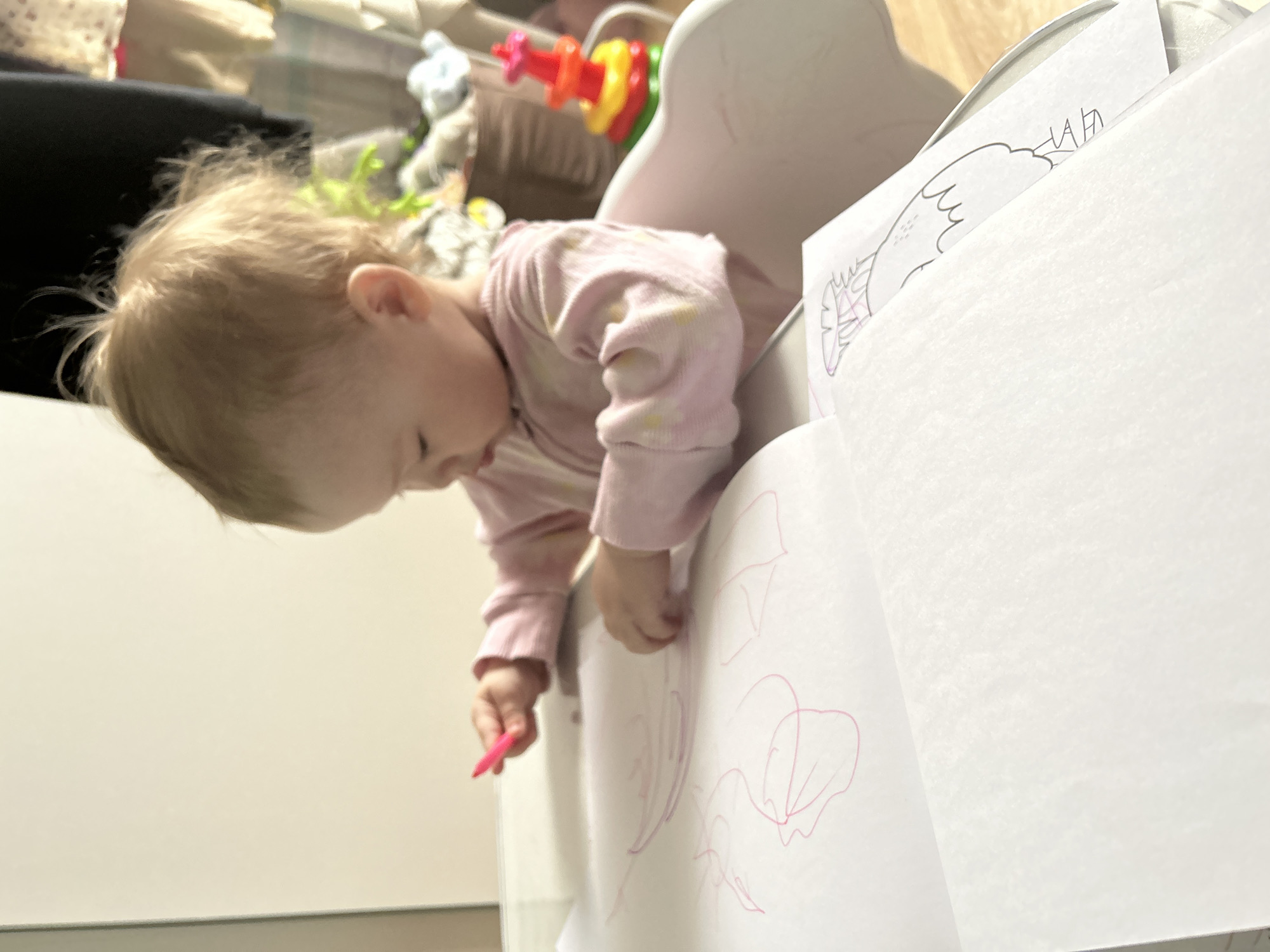 A young child with blonde hair, wearing a pink long-sleeved shirt, is drawing with a pink crayon on sheets of white paper. The child is seated in a white high chair. The drawing appears to be of flowers. The background shows blurred elements of a domestic interior. There is no evidence indicating the location of this image is the city of Chaikovsky, Russia.