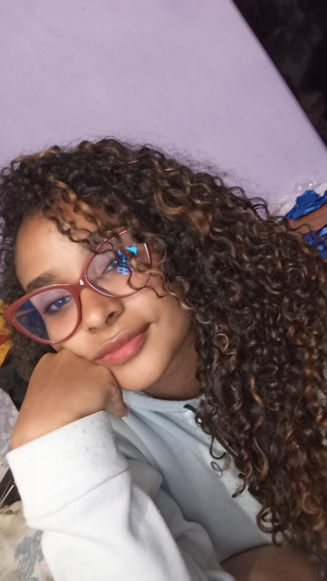 A young person with curly, brown and blonde-highlighted hair and glasses is resting their face on their hand. They are wearing a light blue sweatshirt. The background appears to be a lavender-colored wall or bedding. The location is unspecified in the image, and there is no information to identify the scene as Juan Griego, Venezuela.
