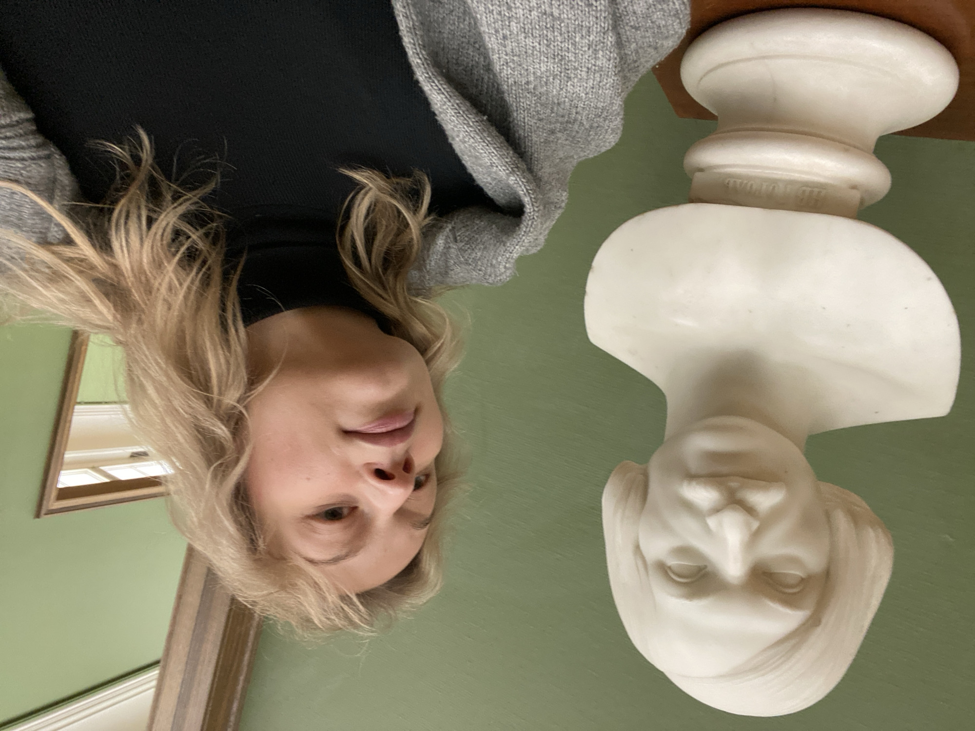 A woman with blonde hair, wearing a black top and a grey cardigan, stands next to a white marble bust. The bust is on a small wooden pedestal. The background is a pale green wall. A framed mirror hangs on the wall. The location appears to be a museum or gallery, possibly in Moscow, Russia.