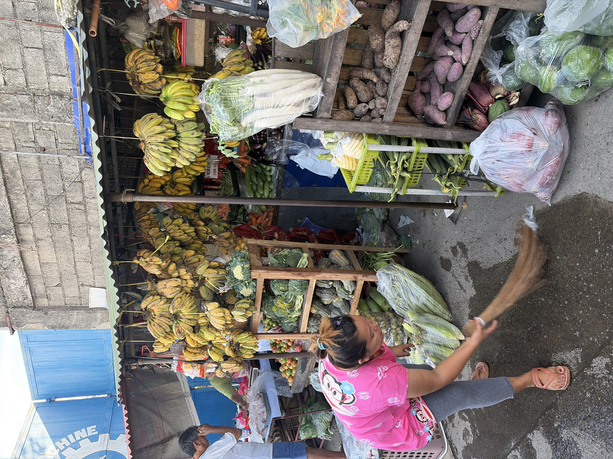 This image depicts an outdoor market scene in Dagupan City, Philippines. A woman in a pink shirt and grey leggings is sweeping the ground with a broom. She wears pink sandals. In front of her are large, clear plastic bags filled with green produce, possibly bananas or plantains. Behind her, a wooden stall displays a variety of fresh produce including bananas hanging in bunches, leafy green vegetables like broccoli and cabbage, and root vegetables such as yams and sweet potatoes. A man in a white t-shirt and blue shorts stands to the left, near a table laden with more fruits and vegetables. The market is set against a backdrop of a brick wall and a blue corrugated metal structure. The ground is paved and appears wet in some areas.