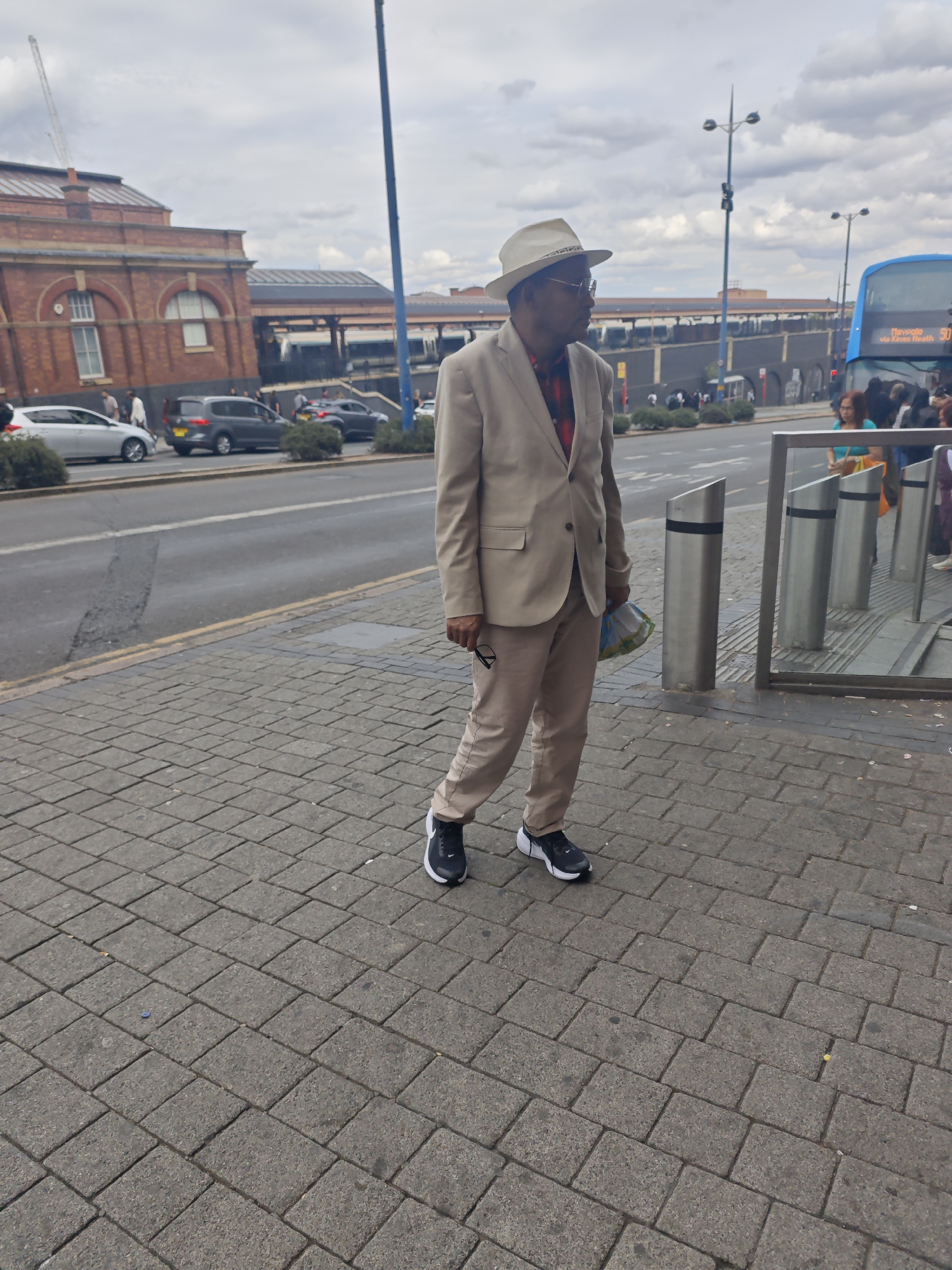 A man, dressed in a tan suit, red and black checkered shirt, white fedora, and black athletic shoes, stands on a paved walkway. He holds a plastic bag in his right hand and eyeglasses in his left. In the background, a blue double-decker bus is visible, along with cars on a street and buildings that suggest a city environment. The bus has text on its front that reads "Heypole via Kines Heath 50". The presence of the bus route information suggests this location is likely in or near Birmingham, United Kingdom, possibly near a bus station or transport hub. The weather appears overcast.