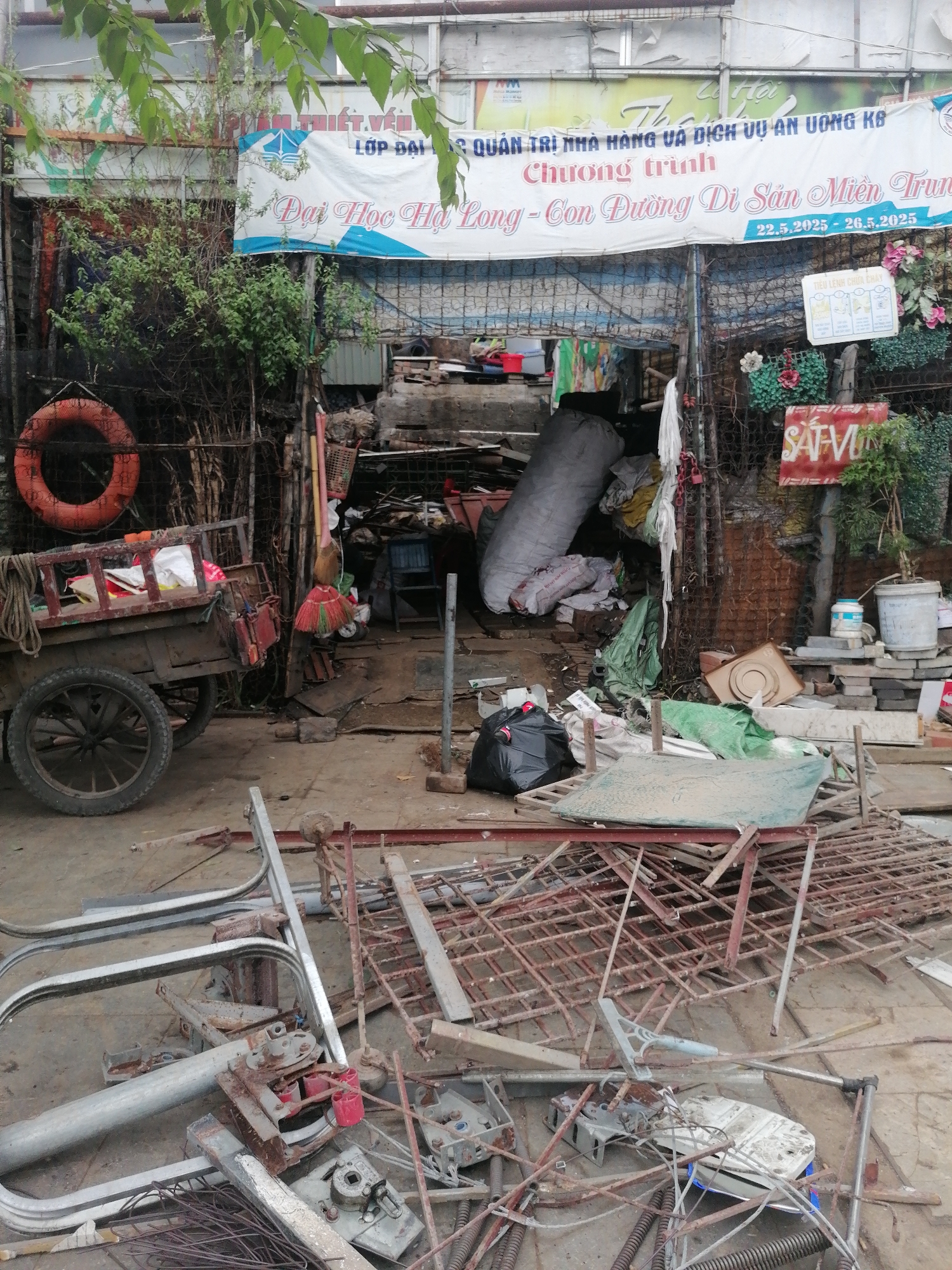 The image depicts a cluttered outdoor scene in Ha Tu Ward, Vietnam. In the foreground, a pile of scrap metal and discarded machinery parts lies scattered on the ground. Behind this, a wooden cart with a wheel is visible, along with a life preserver hanging on a wire fence. The entrance to a building is obscured by piles of refuse, including a large white bag, and various other objects. A banner that reads "Dai Hoc Ha Long - Con Duong Di San Mien Trung" (Ha Long University - The Heritage Road of Central Vietnam) is stretched across the top of the building, with dates "22.5.2025 - 26.5.2025." A sign marked "SAFVUN" is also visible amidst the foliage and debris. The overall environment appears disorganized and neglected.