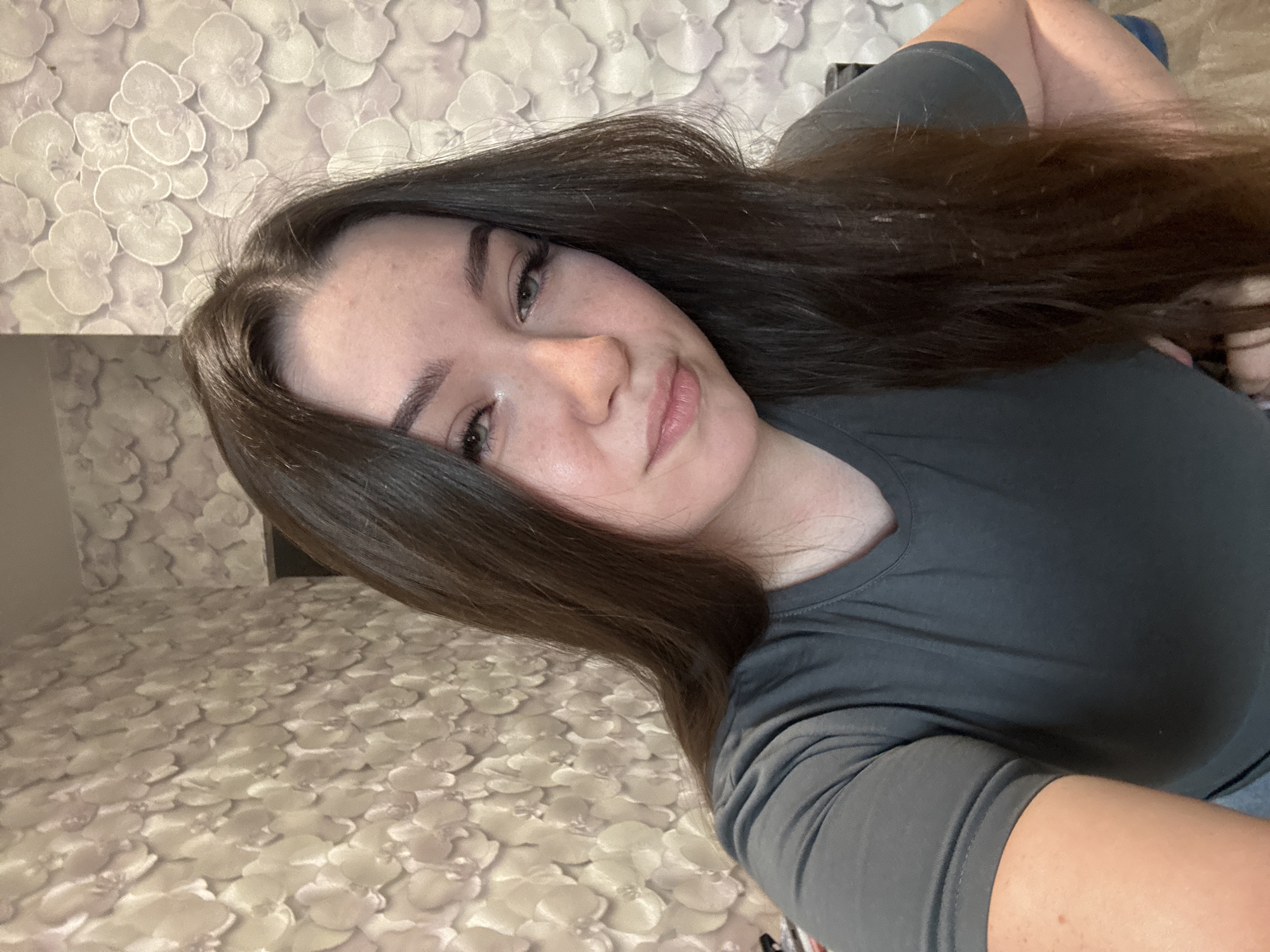 The image is a selfie of a young woman with long, dark brown hair and fair skin. She is wearing a dark gray t-shirt. Her head is tilted slightly to the right, and she is looking towards the camera with a gentle expression. The background consists of floral wallpaper in muted tones of beige and white. The scene appears to be indoors, possibly in a residential setting within Zarechnaya (Gamovskoe S/P), Russia. There are no other individuals or notable objects in the frame besides the woman and the wallpapered wall.