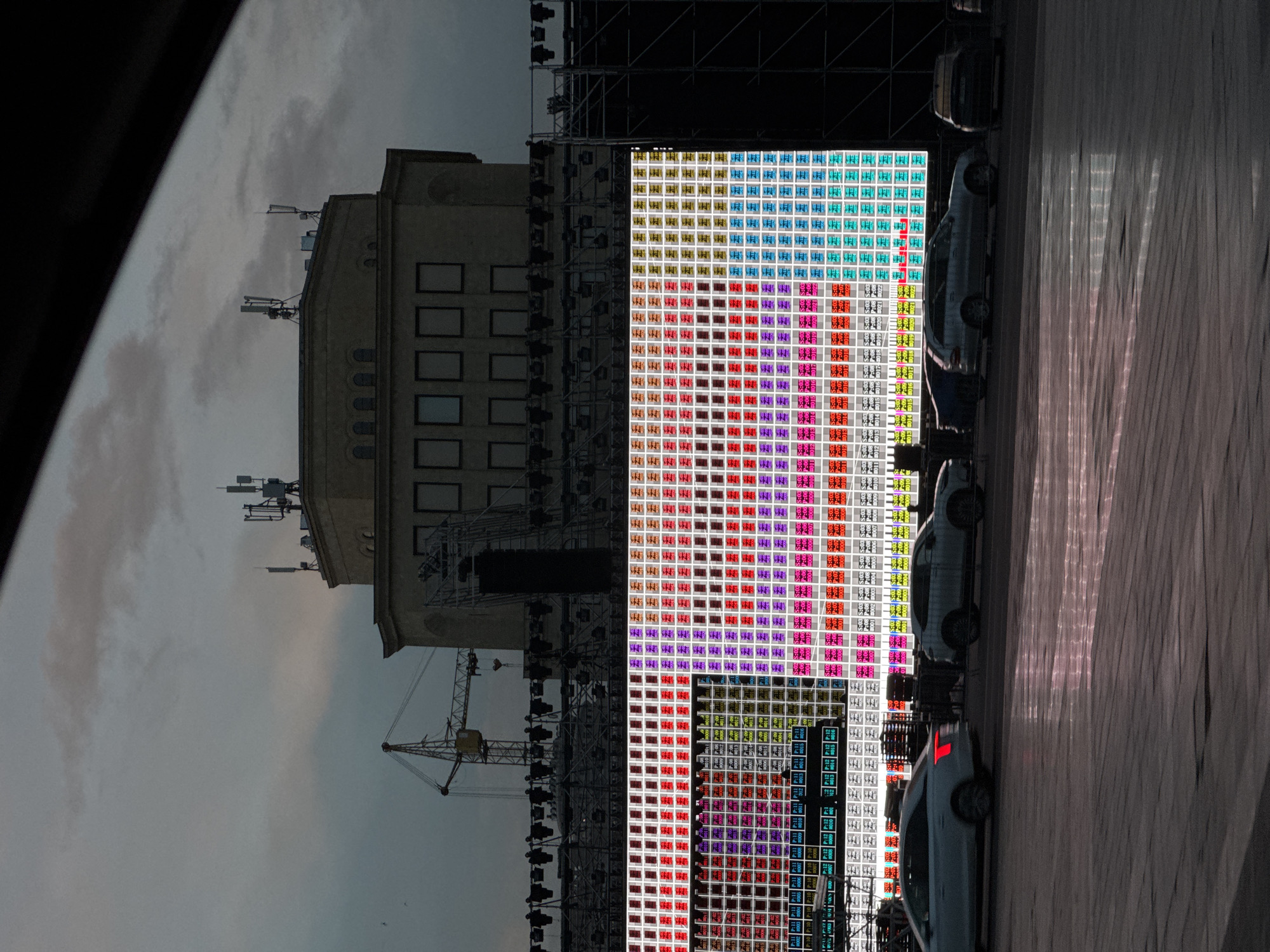The image shows a sideways view of Republic Square in Yerevan, Armenia. In the background is a beige building with multiple windows and telecommunication towers on top. To the right of the building is a large outdoor LED screen displaying a colorful, pixelated pattern. In front of the screen, several cars are parked. The right side of the image features a blurry, grey area, likely a window frame. A construction crane is visible near the building. The sky is cloudy.