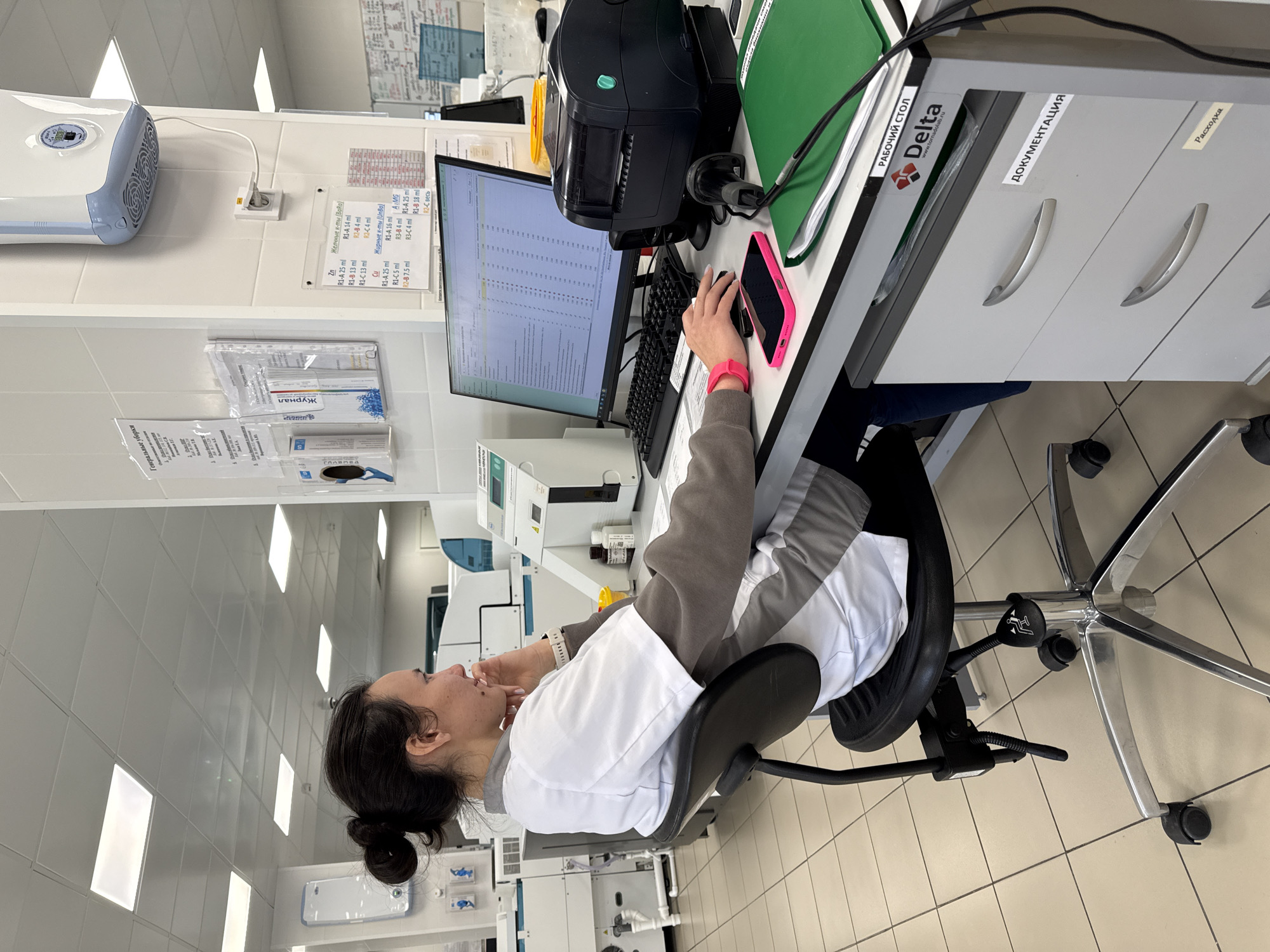 A woman in a white lab coat sits at a workstation in what appears to be a laboratory setting, potentially in Moscow, Russia given the Cyrillic script on signs. She's looking at a computer screen, her hand near her face as if in thought. A pink smartphone rests on the desk near a keyboard. Equipment such as printers and drawers labeled "Documentation" are also present at the workstation. The setting appears sterile, with tile flooring and fluorescent lighting. Various analytical instruments and informational posters line the walls of the laboratory.