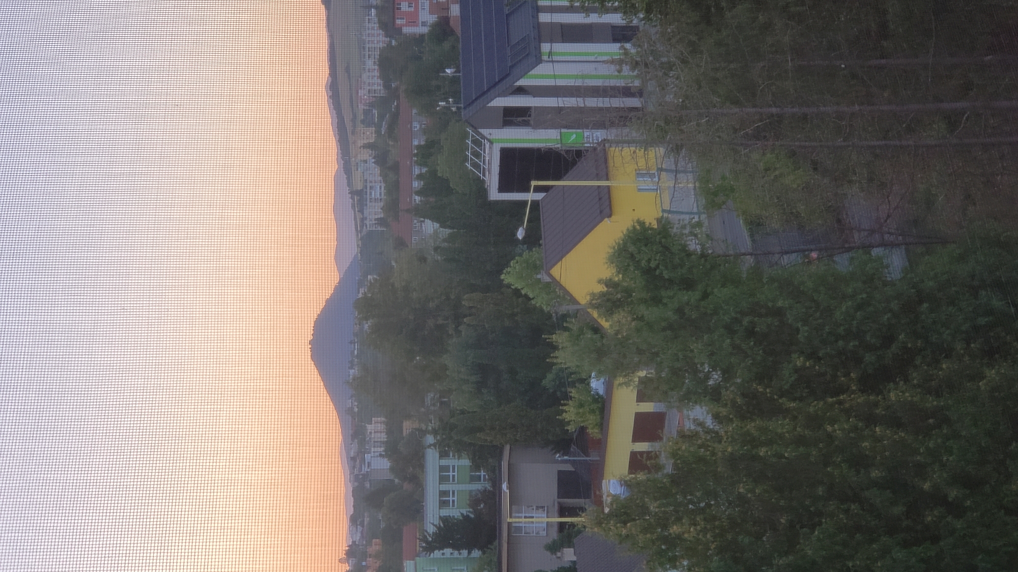 The image captures a view of the city of Prešov, Slovakia, from an elevated vantage point, likely a window with a screen overlaying the scene. The sky is painted with the soft hues of a sunset, transitioning from a warm orange near the horizon to a pale purple at the top. A prominent, rounded hill with a dark, possibly forested, surface dominates the distant landscape, with other rolling hills and a scattered urban environment in the middle ground.
In the foreground and mid-ground, residential and commercial buildings are visible, interspersed with dense greenery. A bright yellow building with a dark roof is situated prominently, featuring garage doors. Adjacent to it, a modern building with white and green vertical stripes and a dark roof is noticeable, equipped with solar panels on its roof. Other structures include a beige building with a white-framed window and a dark roof, and a green building with multiple windows further in the distance. Trees, varying in size and density, are scattered throughout the scene, some appearing as mature evergreens and others as deciduous trees. The overall impression is of a suburban or semi-urban setting during dusk.