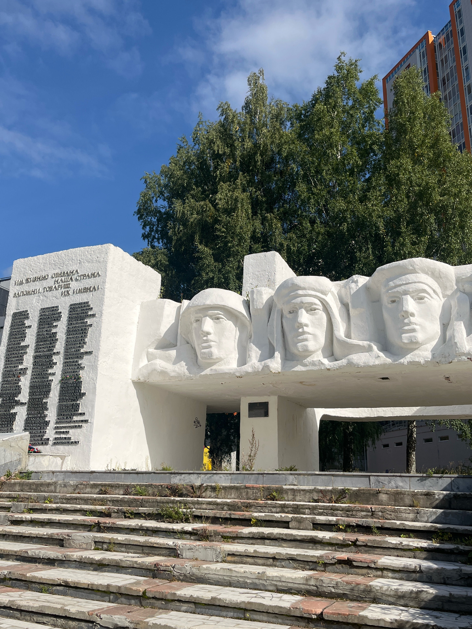 The image shows a monument with three sculpted heads in relief, possibly representing soldiers or historical figures, on a white concrete structure. To the left of the sculpted heads, a white panel features rows of text engraved in black. The monument is situated outdoors, with a clear blue sky visible in the background, punctuated by white clouds and the tops of green trees. A modern building, partly orange and grey, stands to the right, behind the trees. In the foreground, a wide set of concrete stairs, showing signs of wear and overgrown with small plants, leads up to the monument. The overall scene appears to be a public space, likely a memorial or monument in Perm, Russia, given the style of the monument and the Cyrillic text.