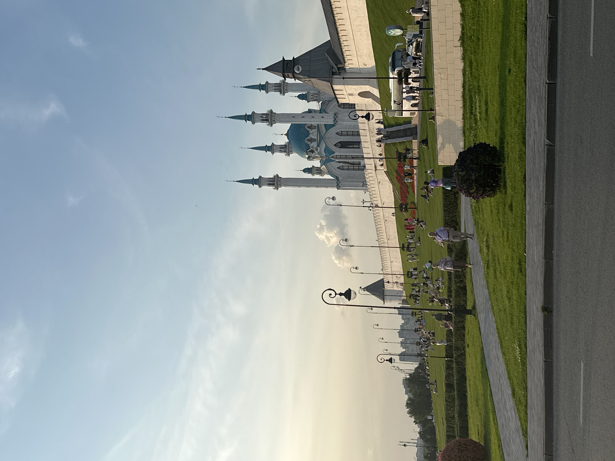 The image shows the Kul Sharif Mosque in Kazan, Russia. The mosque is a prominent landmark with its distinctive turquoise domes and white minarets. It is situated on a grassy hill, with a wall and staircase leading up to the mosque. There are people visible in the foreground and on the hill, some are sitting on the grass, while others are walking. A bus and cars are parked near the base of the hill. The sky is blue with some scattered clouds, and the overall lighting suggests it is late afternoon or early evening. The scene depicts a public space where people are enjoying the surroundings of the mosque.